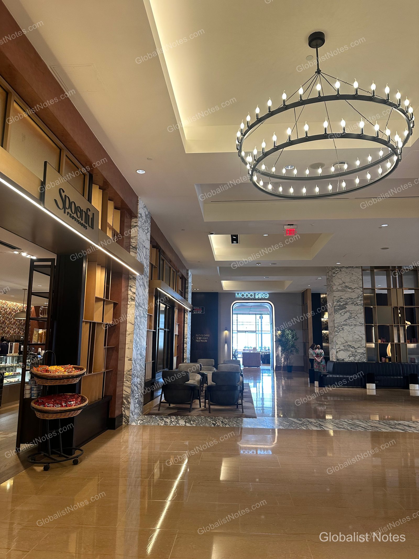 Lobby of the Grand Hyatt San Antonio River Walk, featuring the Spoonful Provisions shop on the left and a large circular chandelier hanging from the ceiling. The space is open and expansive with marble floors and modern furniture.