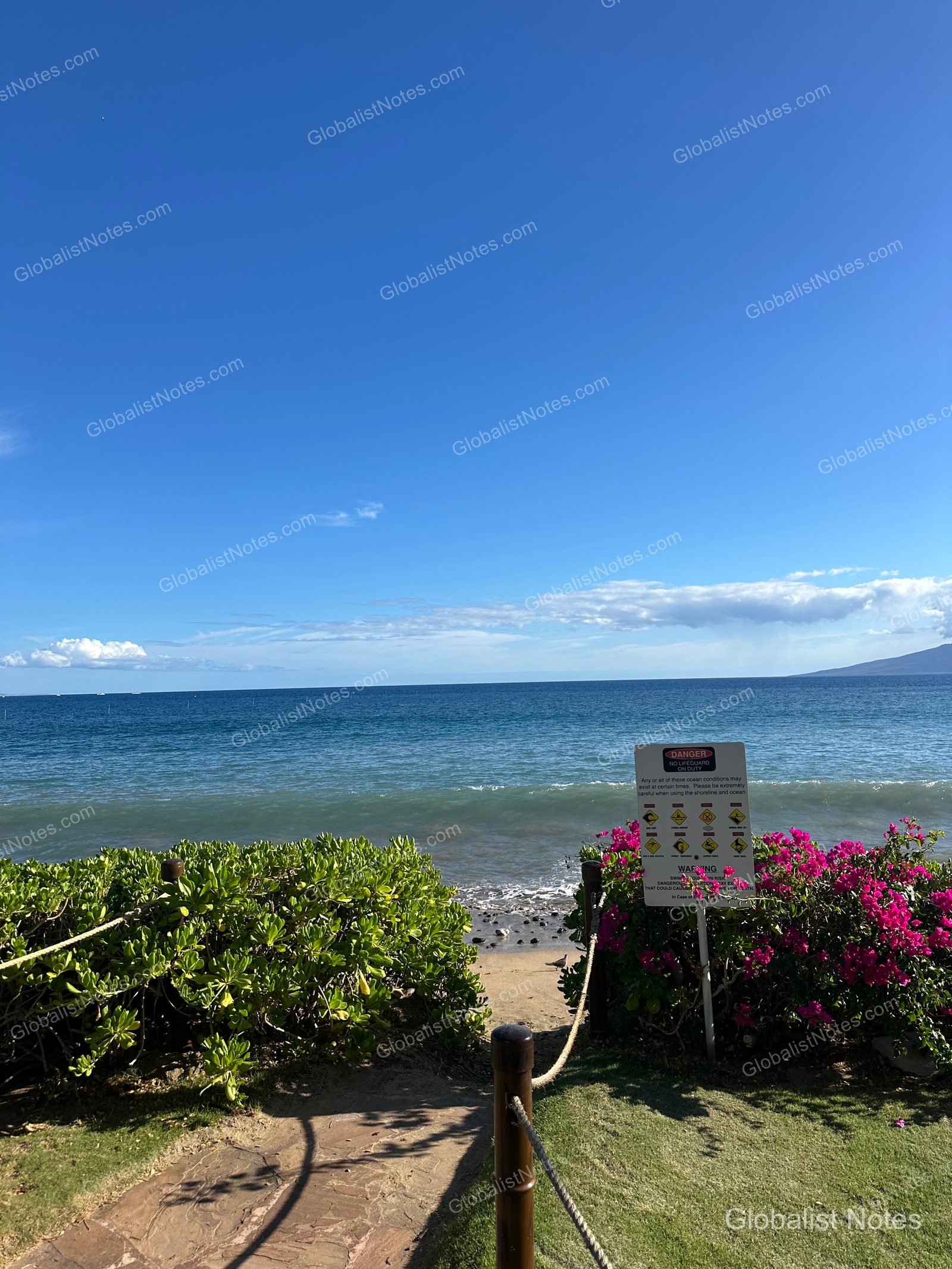 A safety warning sign stands at the end of the path leading to the beach at Hyatt Regency Maui Resort and Spa, with the blue ocean and clear sky in the background.