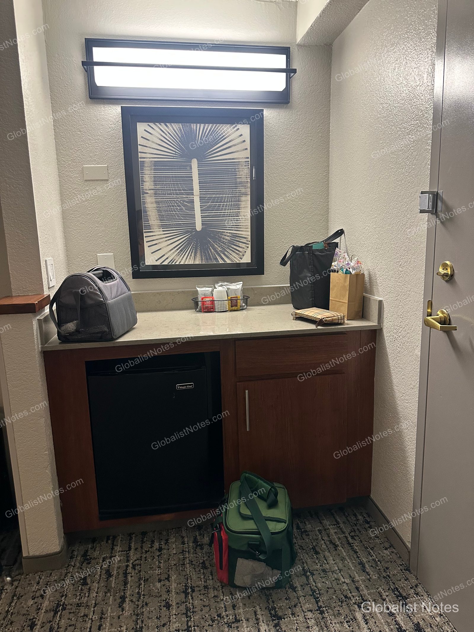 Hyatt Place El Paso Airport Junior Suite 的迷你吧區域。深色木質櫃體上放置了小型冰箱與咖啡機，牆面掛有抽象畫，地面則放著旅行行李箱。