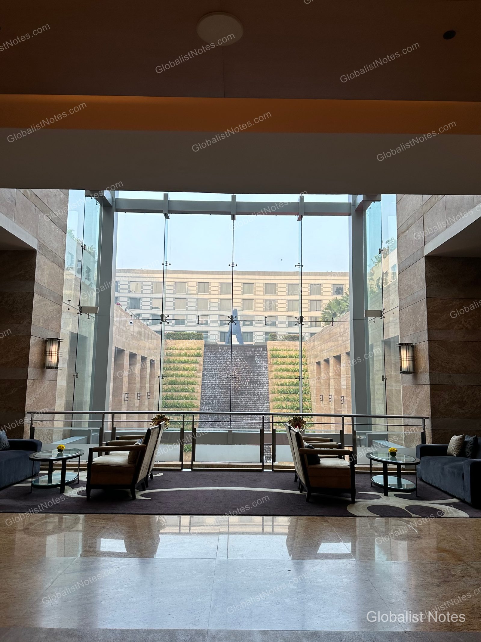 A view through floor-to-ceiling lobby windows at Grand Hyatt Mumbai, overlooking a tiered waterfall and garden. Symmetrical sofas and tables flank the window, set against polished marble floors.
