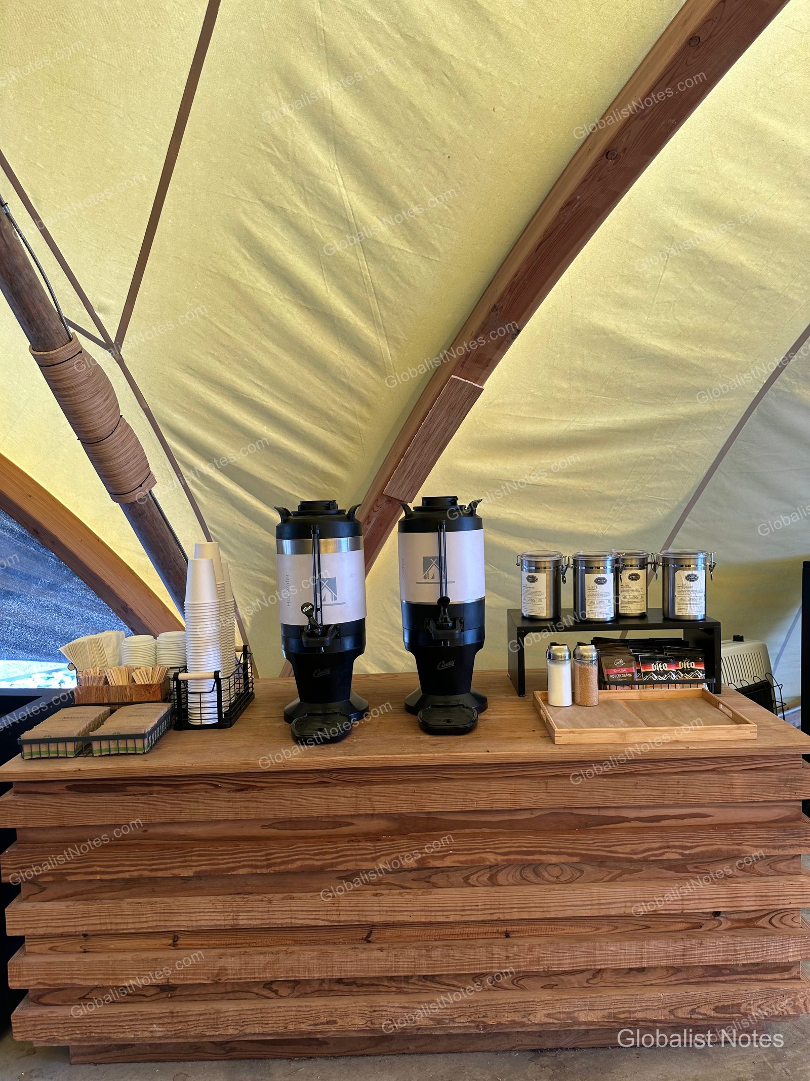 テント内の木製テーブルに置かれたセルフサービスのドリンクステーション。コーヒーサーバー、湯沸かし器、紙コップ、ティーバッグ、砂糖などが並んでいる。