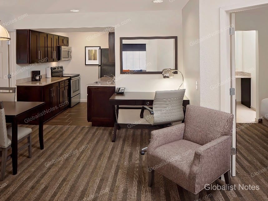 Hyatt House Scottsdale/Old Townのワンベッドルームスイート。ダークウッドのキャビネットとステンレス家電を備えたキッチン、ダイニングテーブル、デスクが配置され、右側には寝室へ続くドアが見えます。
