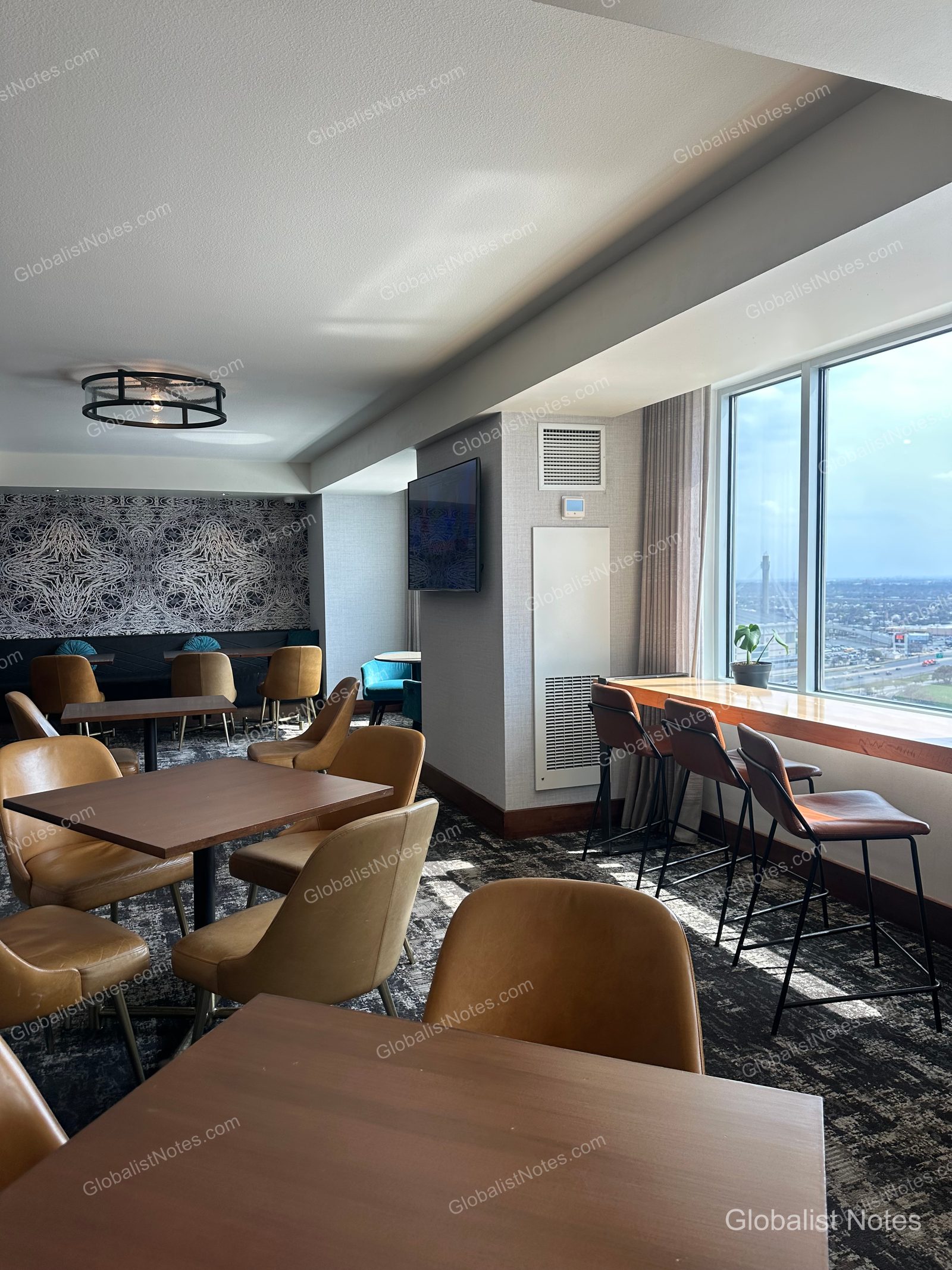 Interior of the lounge at the Grand Hyatt San Antonio River Walk, showing high bar tables and chairs by the window, with square tables and leather chairs in the center. The walls feature geometric black-and-white wallpaper, and the windows offer a view of downtown San Antonio.