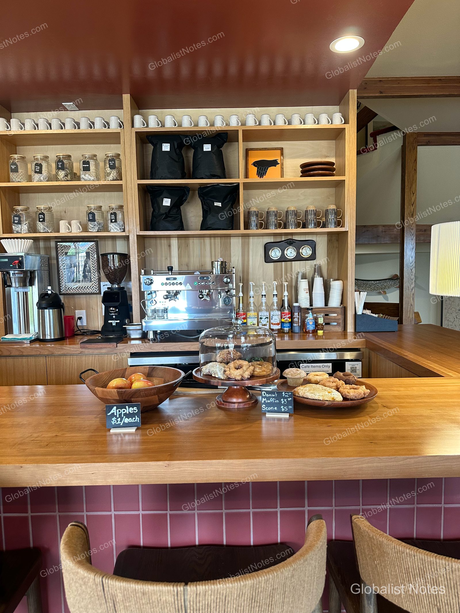 Wanderling Buffaloのカフェカウンター。木製の棚にコーヒーマシンやマグカップ、ガラス瓶が整然と並び、手前のテーブルにはリンゴのボウル、ドーナツ、マフィン、スコーンが置かれています。
