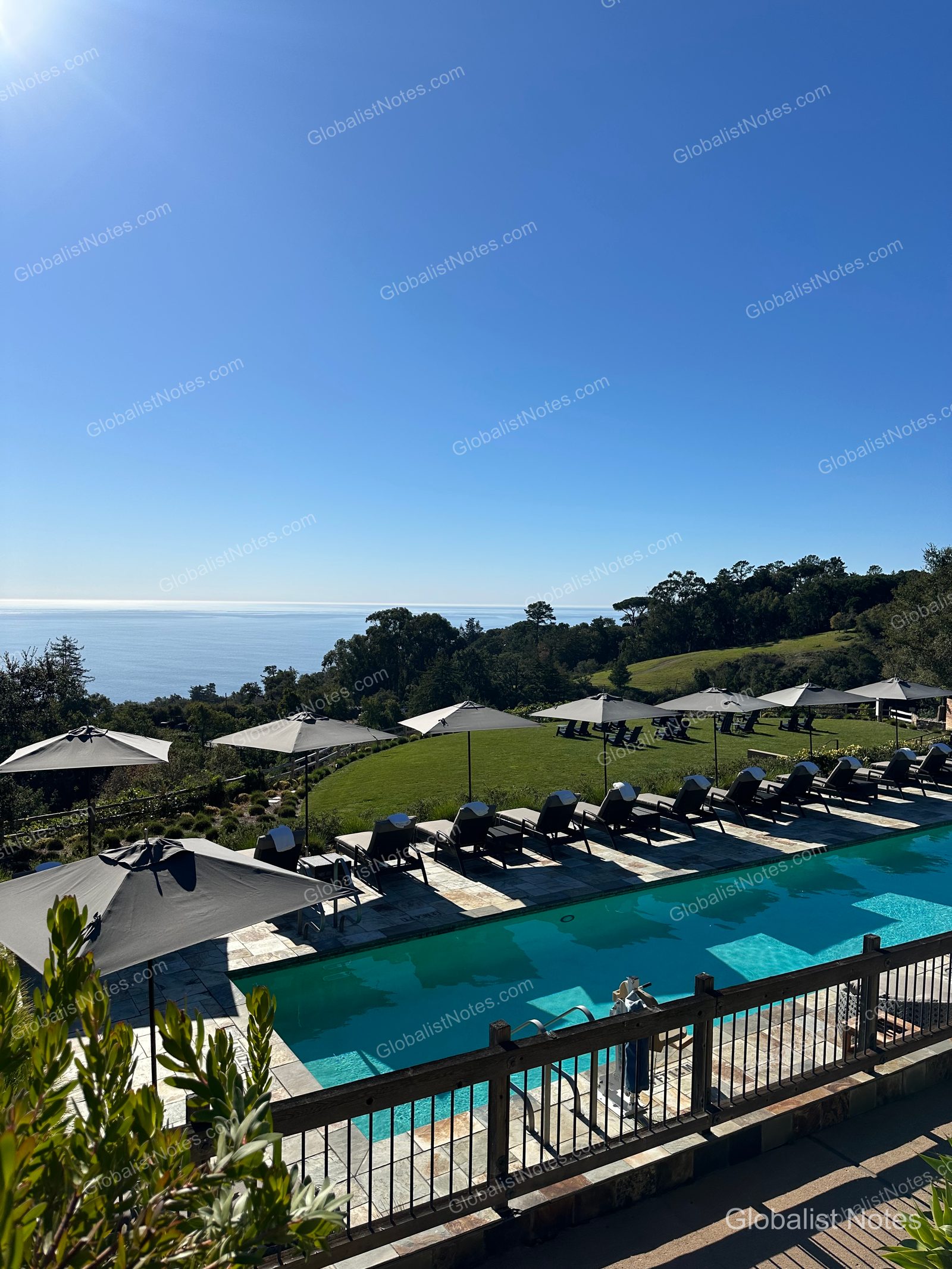 Outdoor pool view at Alila Ventana Big Sur overlooking the Pacific Ocean. Gray umbrellas and sun loungers are lined up along the pool deck, with green lawns and trees in the background.