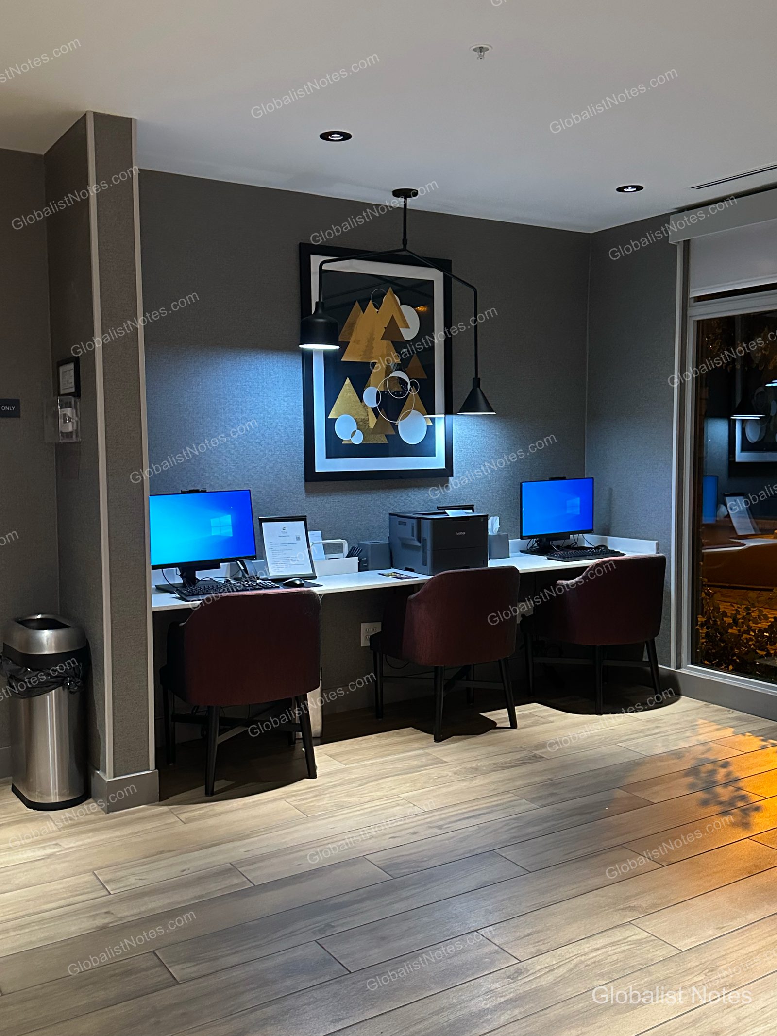 Hyatt Place Flagstaffのビジネスセンター。ダークトーンの壁に幾何学的な山の絵が飾られ、コンピュータ2台とプリンターを備えたデスクが設置されています。