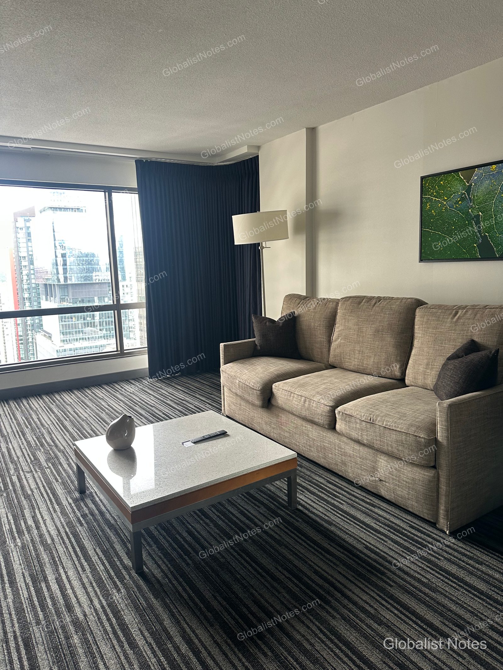 Living area of a Deluxe Suite at Hyatt Regency Vancouver, featuring a beige three-seater sofa, a square marble-topped coffee table, and a floor lamp. Floor-to-ceiling windows offer views of downtown Vancouver high-rises, and the room is finished with striped carpeting.