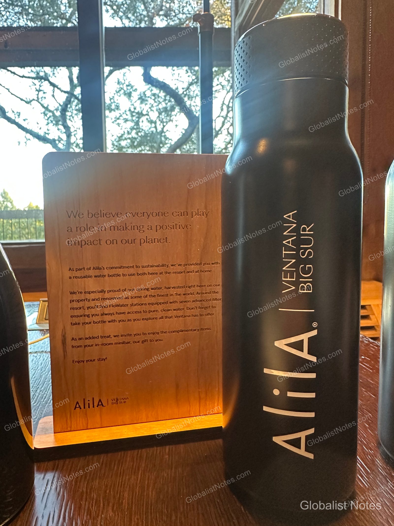 A black reusable water bottle and a wooden sustainability notice placed on the window-side table inside an Alila Ventana Big Sur guest room.