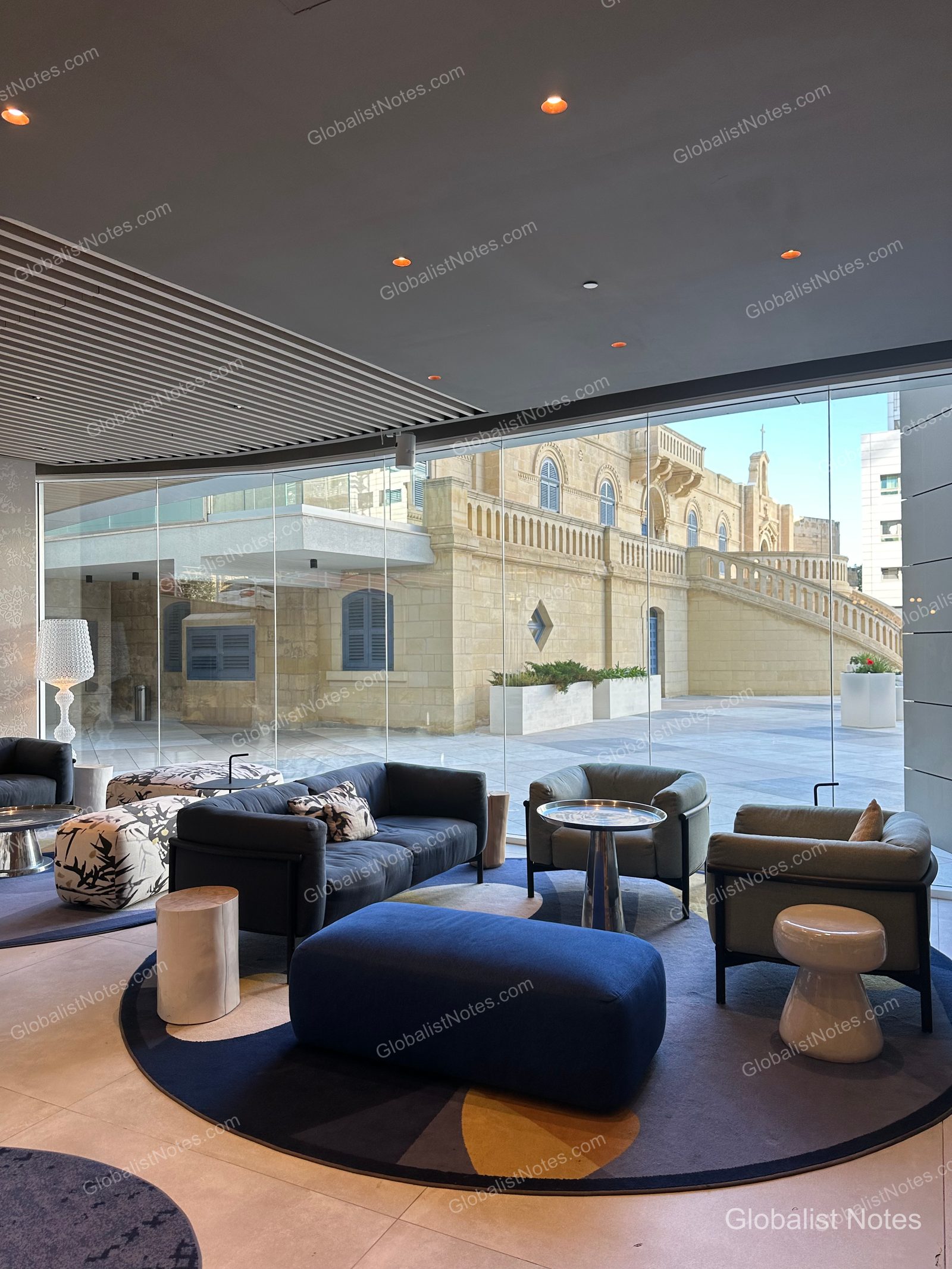 The lobby lounge at Hyatt Regency Malta in St. Julian's. Floor-to-ceiling windows frame the historic stone architecture of St. Augustine's Church, while the interior features deep blue sofas, modern armchairs, and circular rugs.