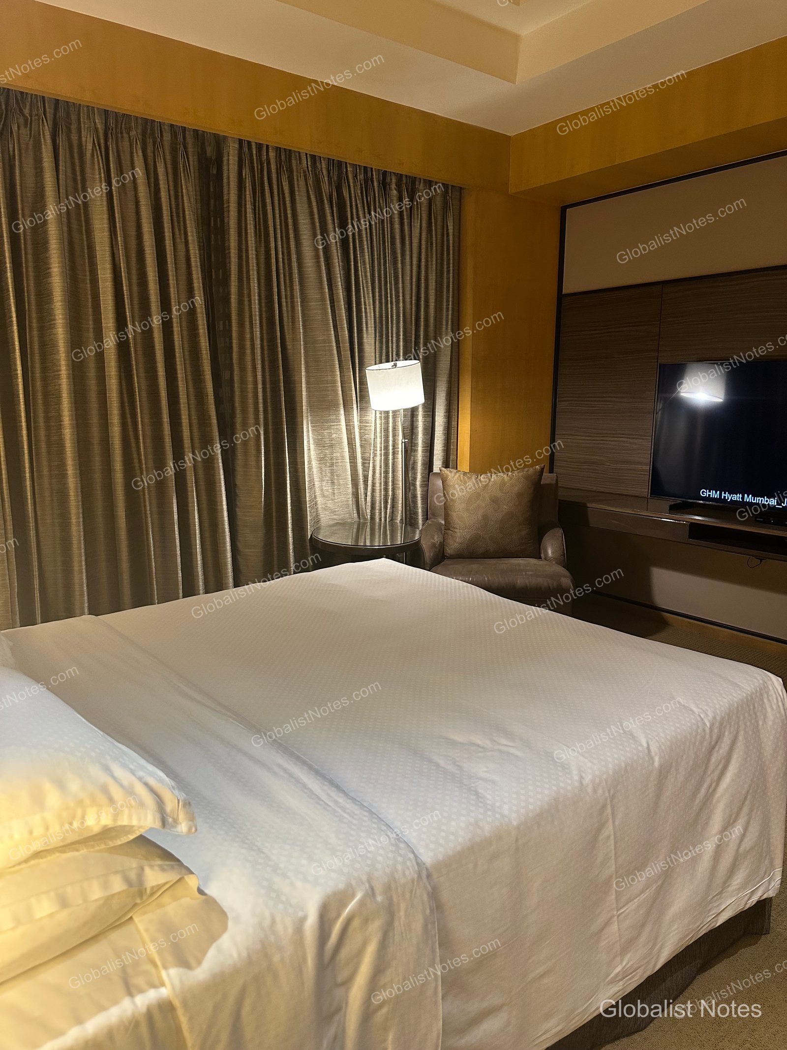 Interior of a Veranda Suite King at Grand Hyatt Mumbai Hotel and Residences, showing a king bed with white linens, an armchair, lighting, and a wall-mounted TV. The windows are dressed in dark brown curtains.