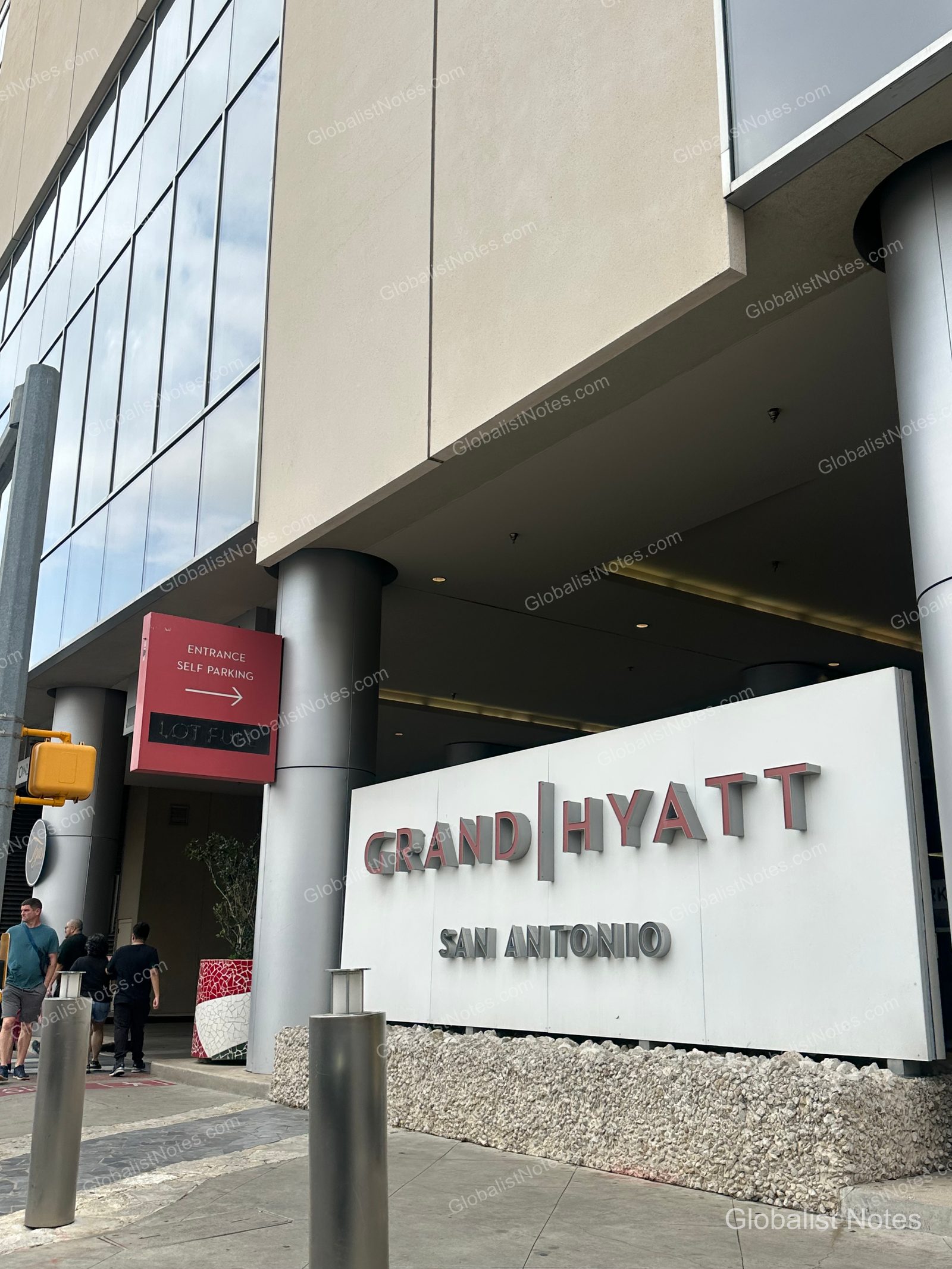 Exterior view of the Grand Hyatt San Antonio River Walk; the hotel name is prominently displayed on the building facade, with a red sign indicating the self-parking entrance and pedestrians walking toward the main entrance.