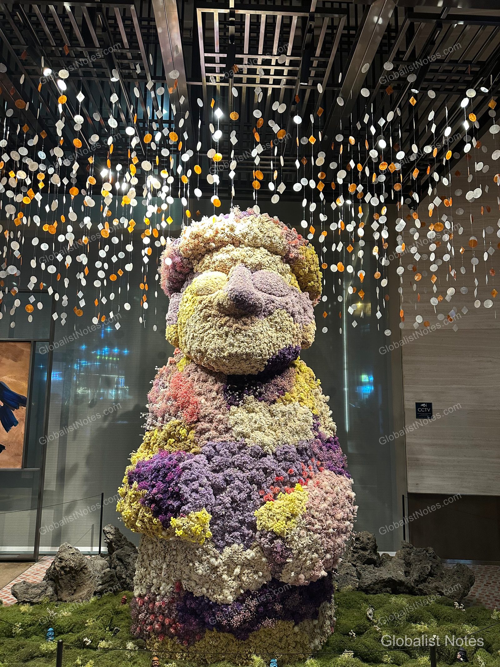 A large Dol Hareubang sculpture located in the center of the Grand Hyatt Jeju lobby. The sculpture is covered in colorful flowers, with numerous circular ornaments hanging from the ceiling above it under bright lighting, and the base is decorated with moss and basalt rocks.