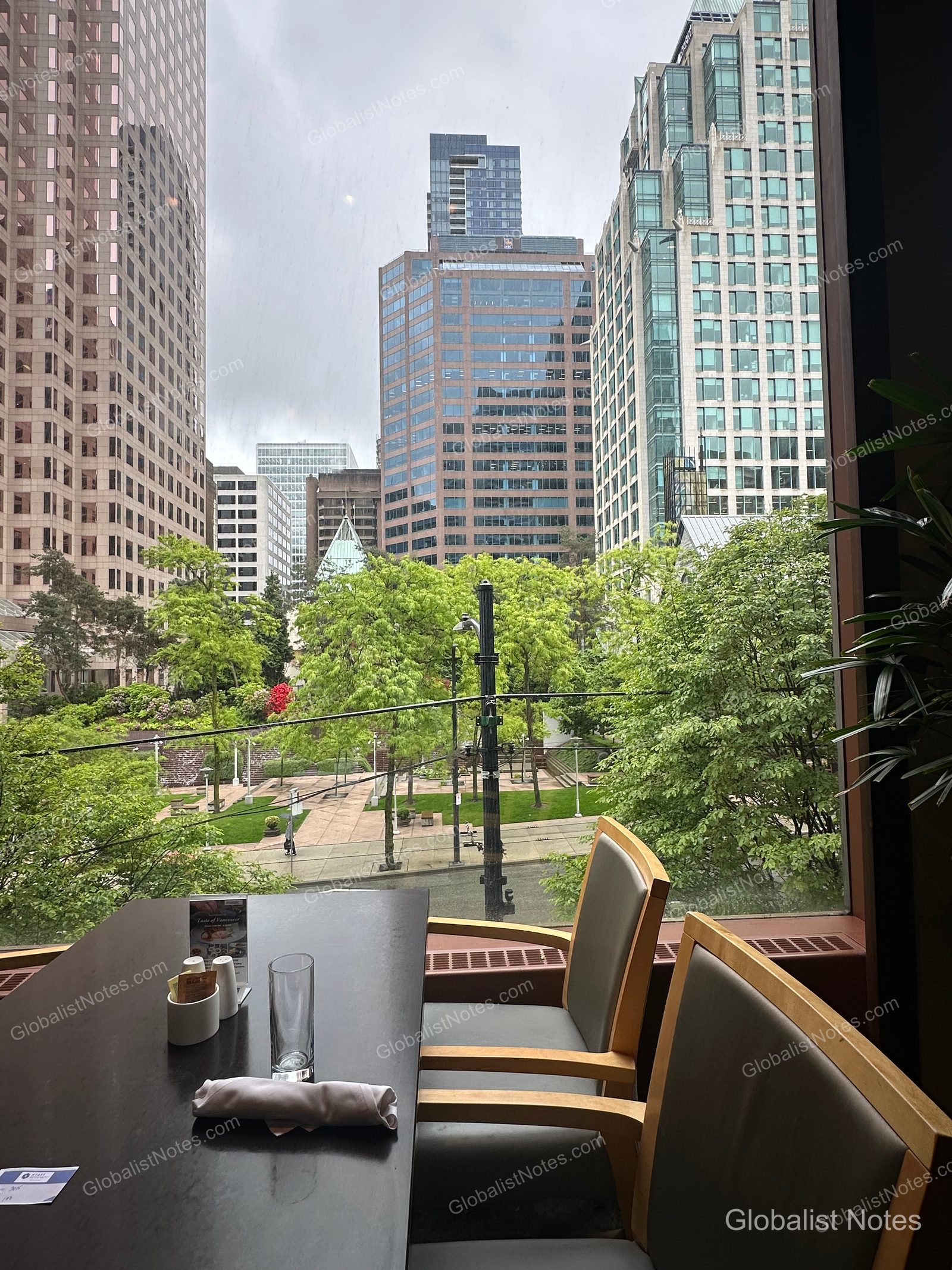 City view from a window seat in the Hyatt Regency Vancouver restaurant. An empty glass and napkin sit on the table, with a view of a lush green park and surrounding high-rise buildings under a cloudy sky.