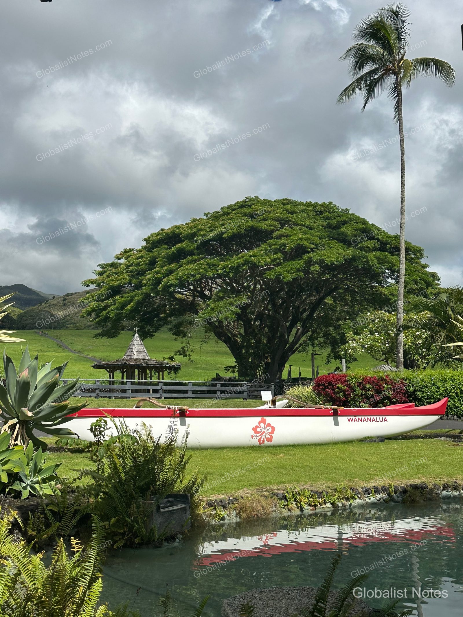 A traditional canoe with red patterns sits on the green lawn at Hana-Maui Resort, with large trees, palm trees, and a small pavilion visible in the background under a cloudy sky.
