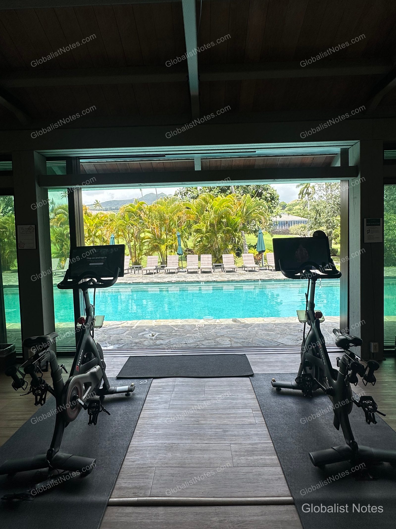 View from inside the fitness center at Hana-Maui Resort. Two indoor cycling bikes are positioned facing the open windows, which overlook the outdoor pool, lush gardens, and rows of sun loungers.