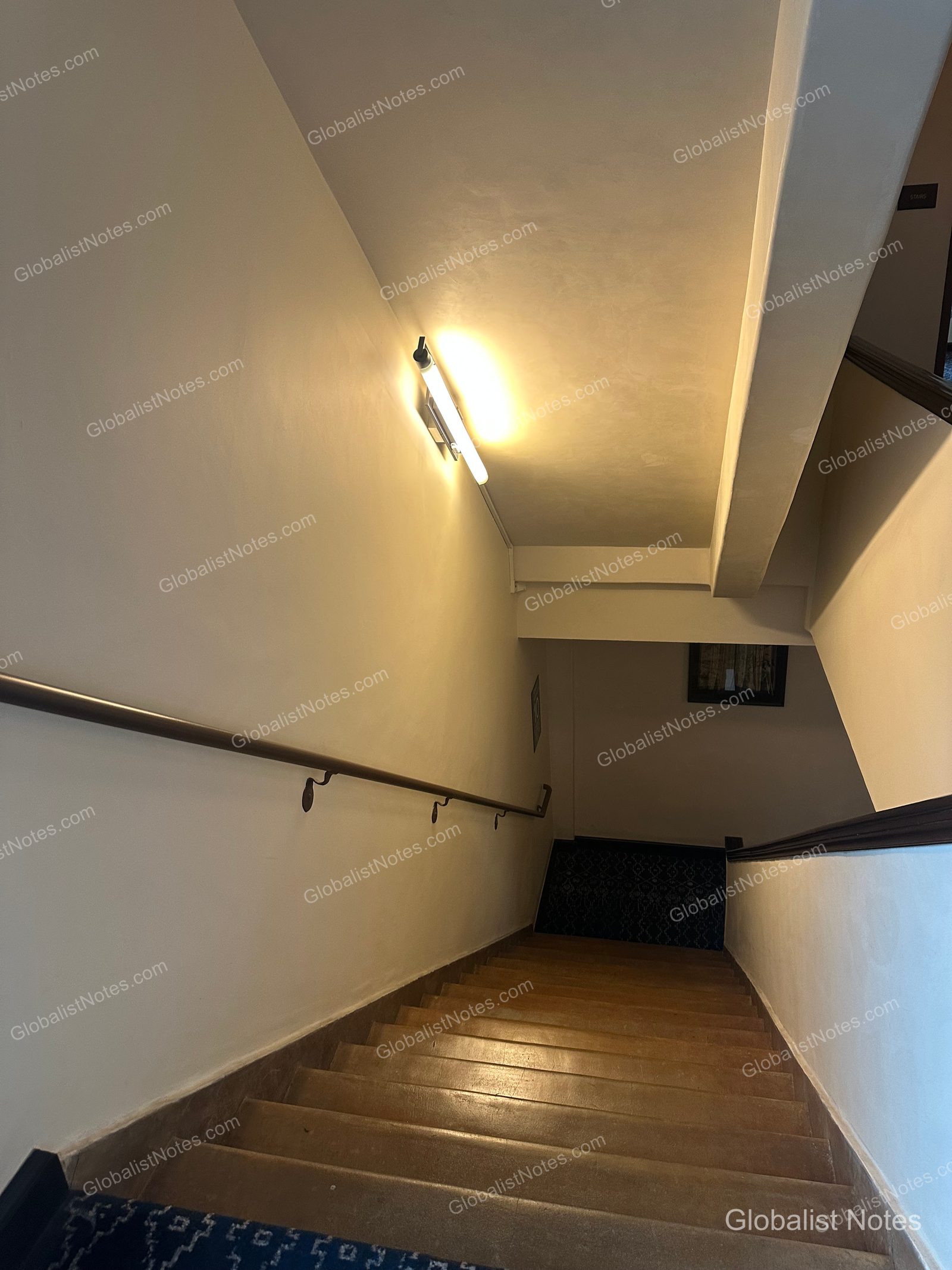 The staircase at Hotel Figueroa in Los Angeles, featuring elongated lighting on bright walls and dark railings along the stairs. The staircase has a muted-tone floor and leads down to the lower level.