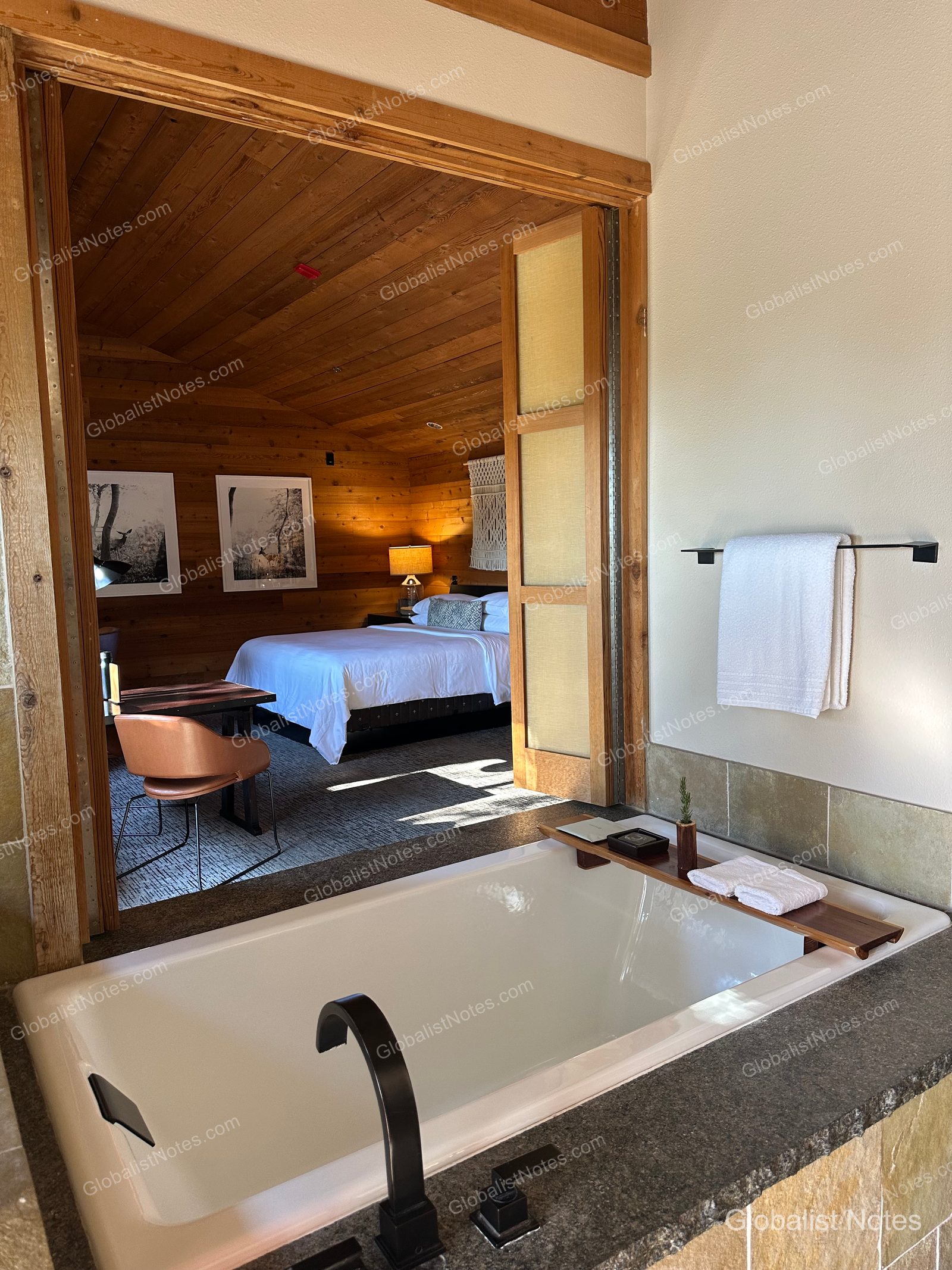 View of the bedroom from the bathroom of a Junior Suite at Alila Ventana Big Sur, showing a bathtub in the foreground and the bedroom area visible through sliding doors with wood-paneled walls and warm lighting.