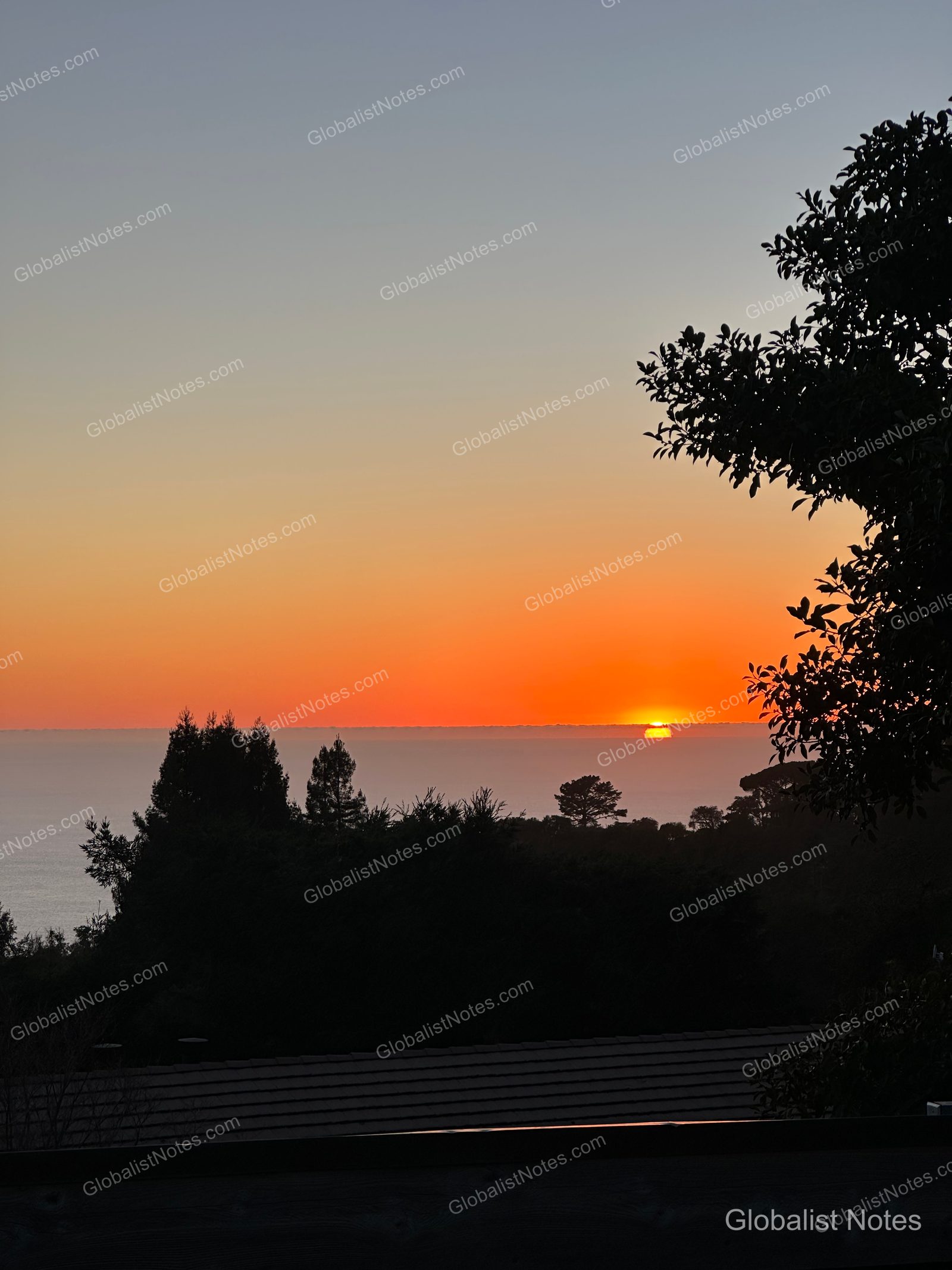 Sunset view from Alila Ventana Big Sur in Big Sur, USA. The sun sets over the horizon, turning the sky orange and purple, with the silhouettes of trees and a portion of a building roof in the foreground.