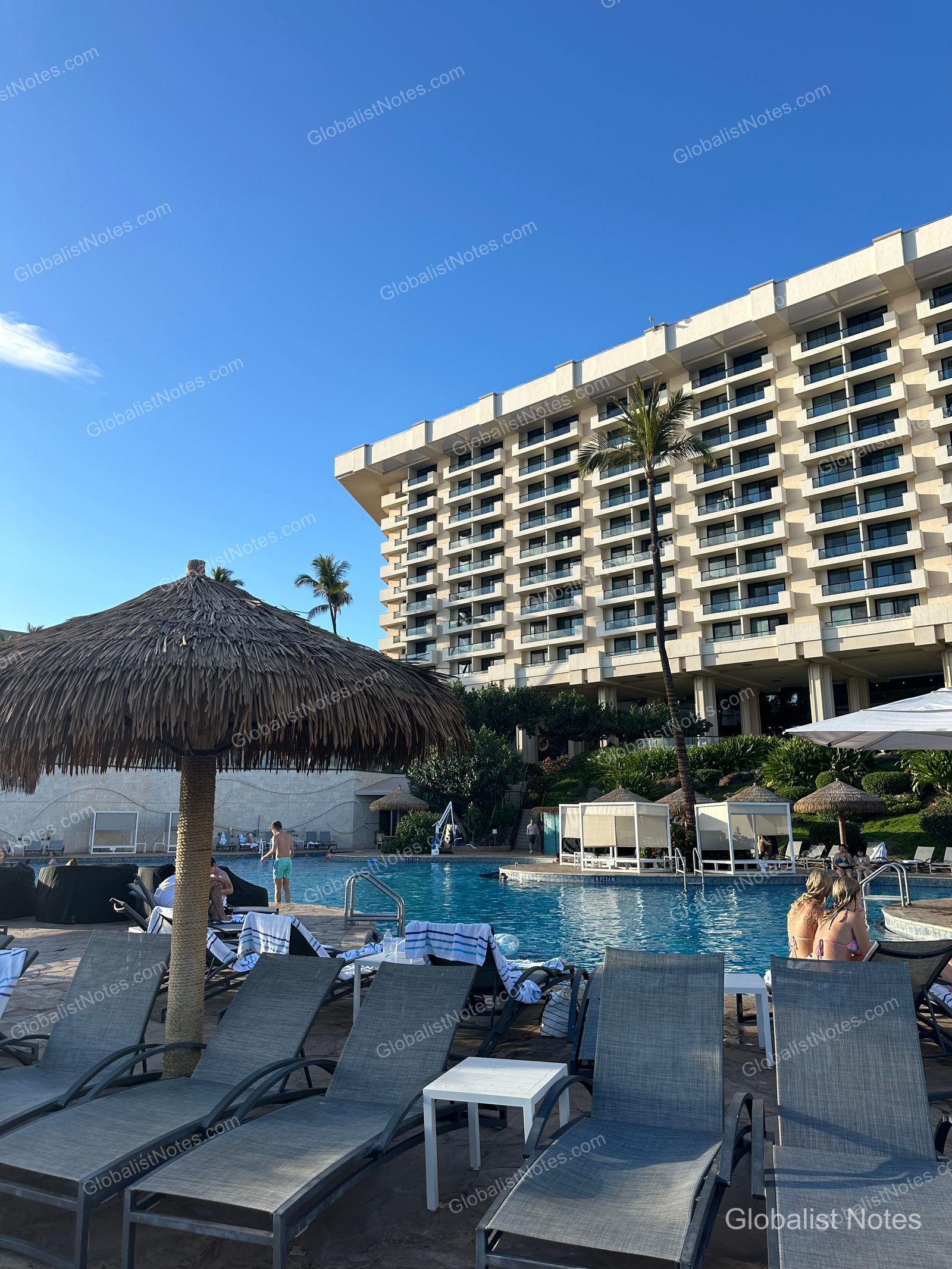 Outdoor pool view at Hyatt Regency Maui Resort and Spa in Lahaina, Hawaii. Rows of thatched umbrellas and sun loungers line the pool, with the resort's guest room towers in the background and guests swimming under a blue sky.