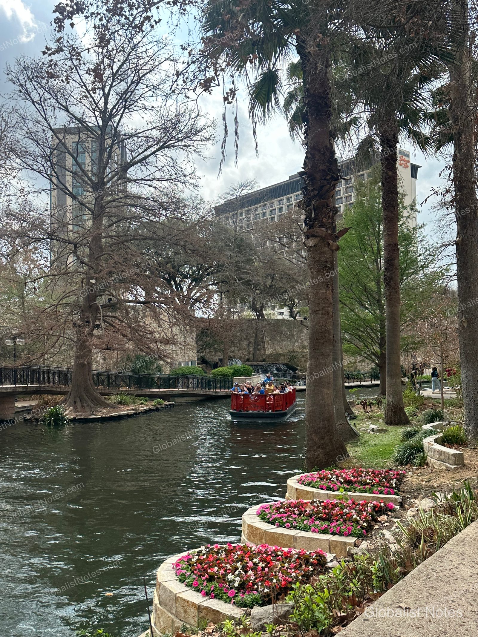 A tourist boat cruises along the San Antonio River Walk canal, framed by lush trees and flower beds, with the Grand Hyatt San Antonio River Walk building visible in the background.