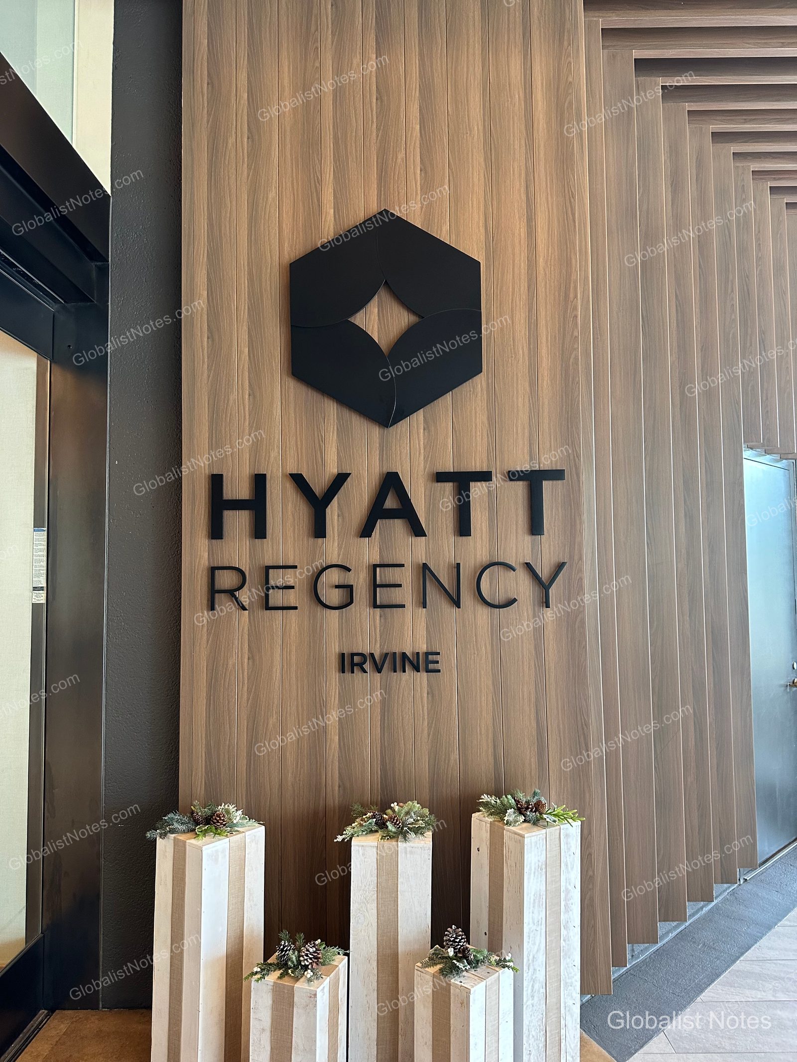 Hyatt Regency Irvine 大廳牆面上的黑色品牌標誌與飯店名稱。背景為垂直木條裝飾，下方擺放著裝飾有松果與樹葉的木柱，營造出冬季氛圍。