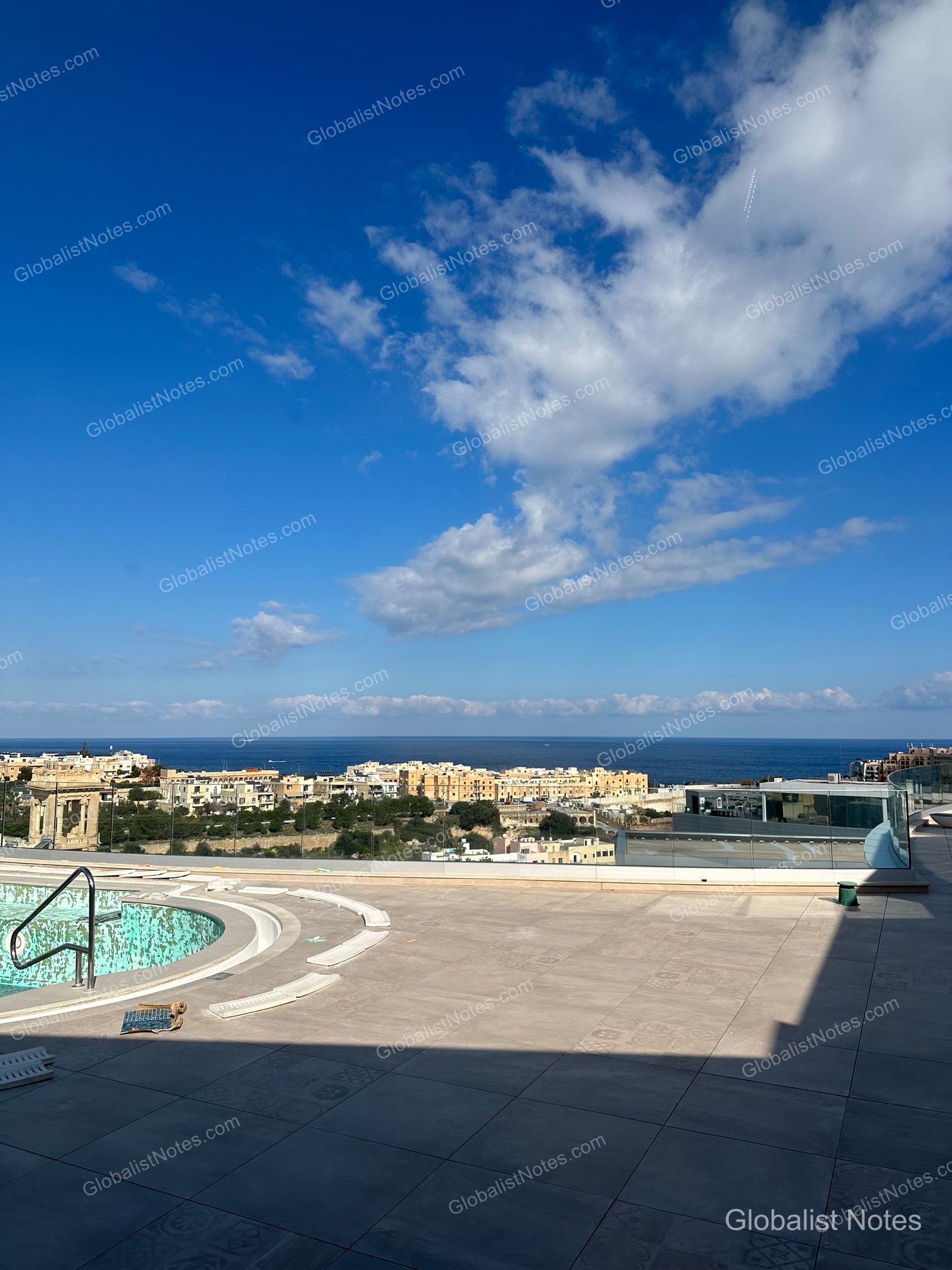 A view from the rooftop pool at Hyatt Regency Malta, looking out over the St. Julian's cityscape and the Mediterranean Sea beyond the glass railing.