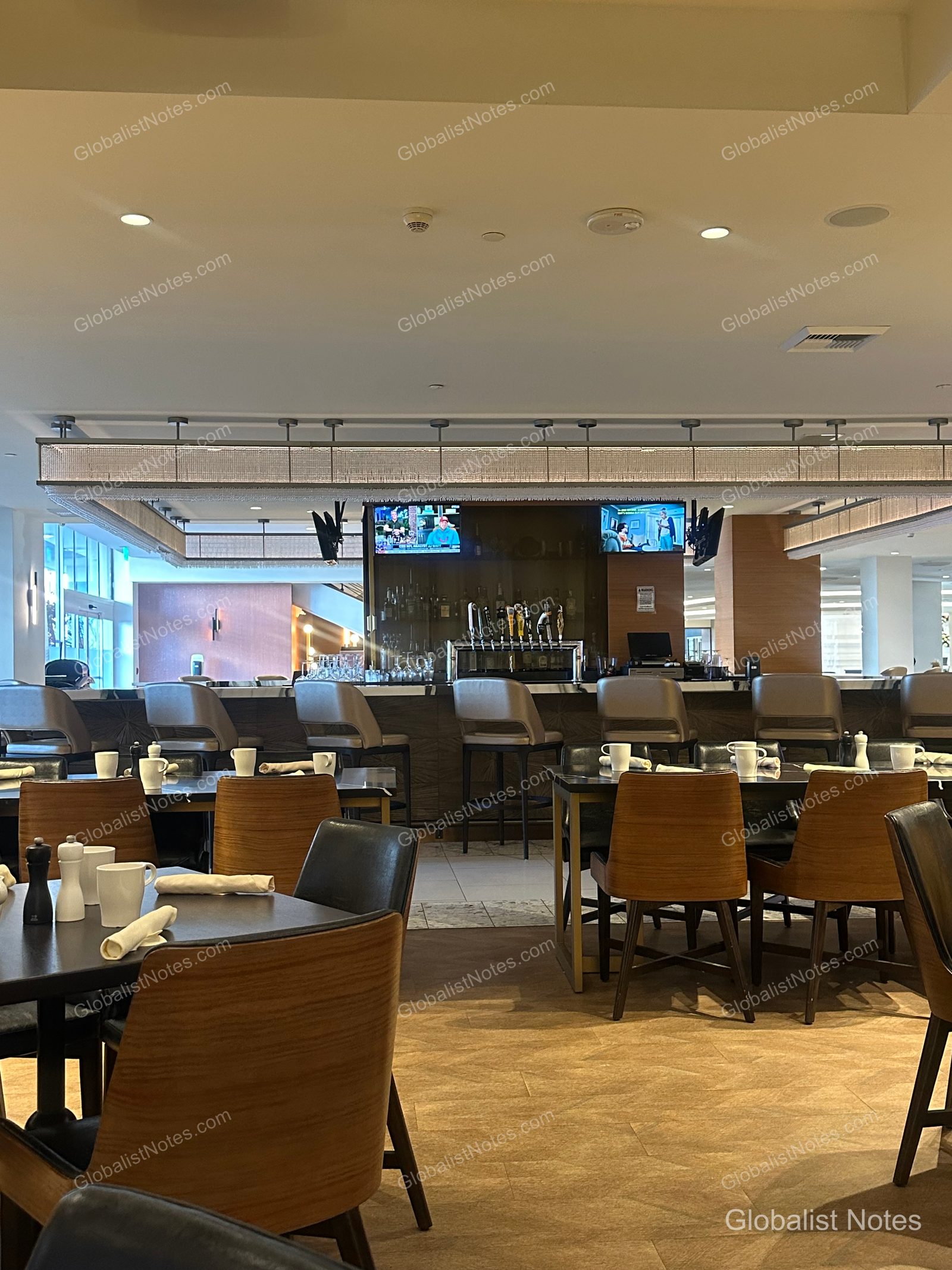 Interior view of the breakfast restaurant at Hyatt Regency Los Angeles International Airport. The space features neatly arranged wood-toned tables and chairs, with a modern dining area in the background equipped with a bar counter and TVs.