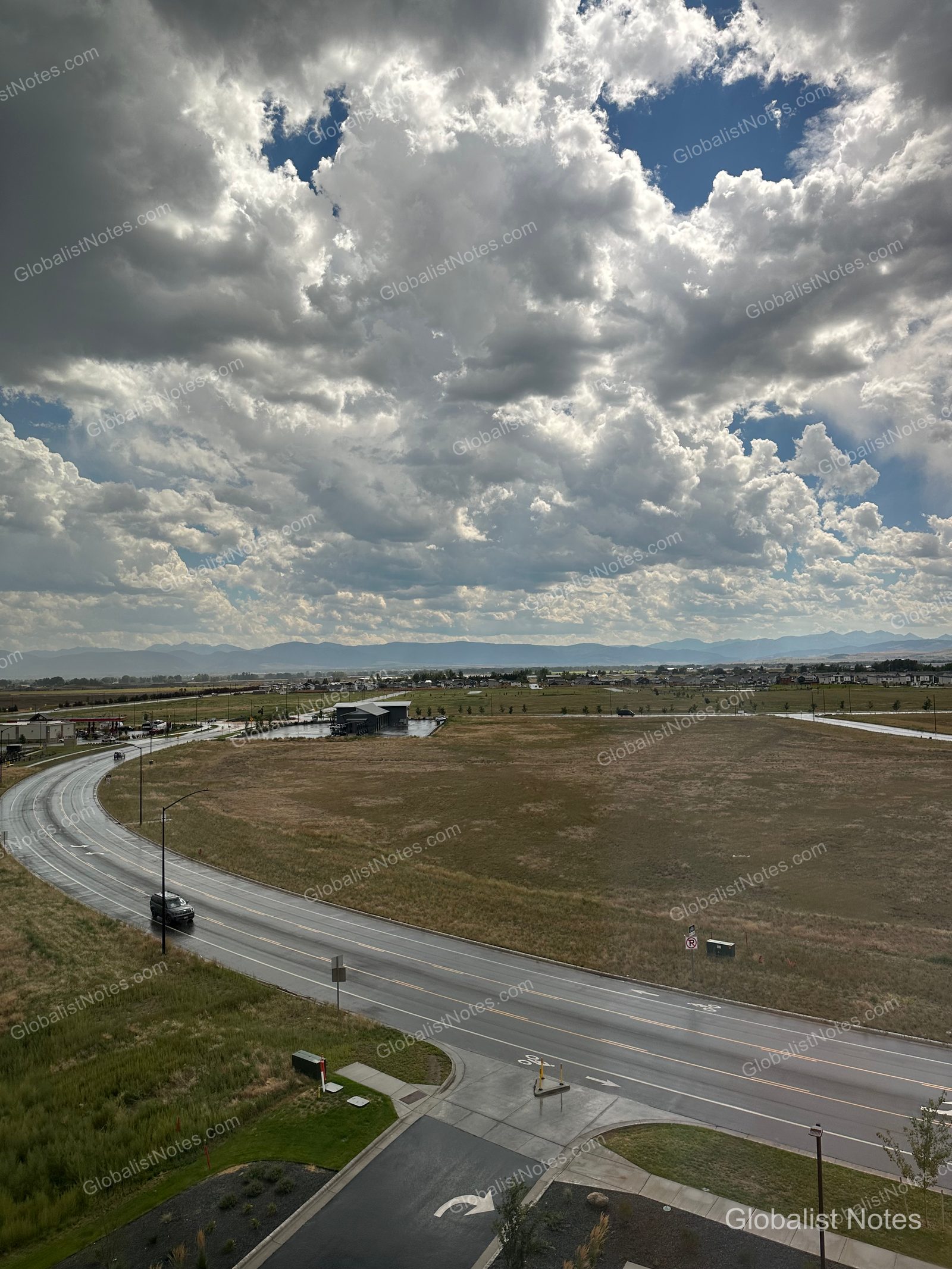 Hyatt Place Bozeman Yellowstone Airportのジュニアスイートルームから望むベルグレードの広大な景色。雨に濡れた道路が曲線を描き、曇り空の下に平原と遠くの山脈が広がっています。