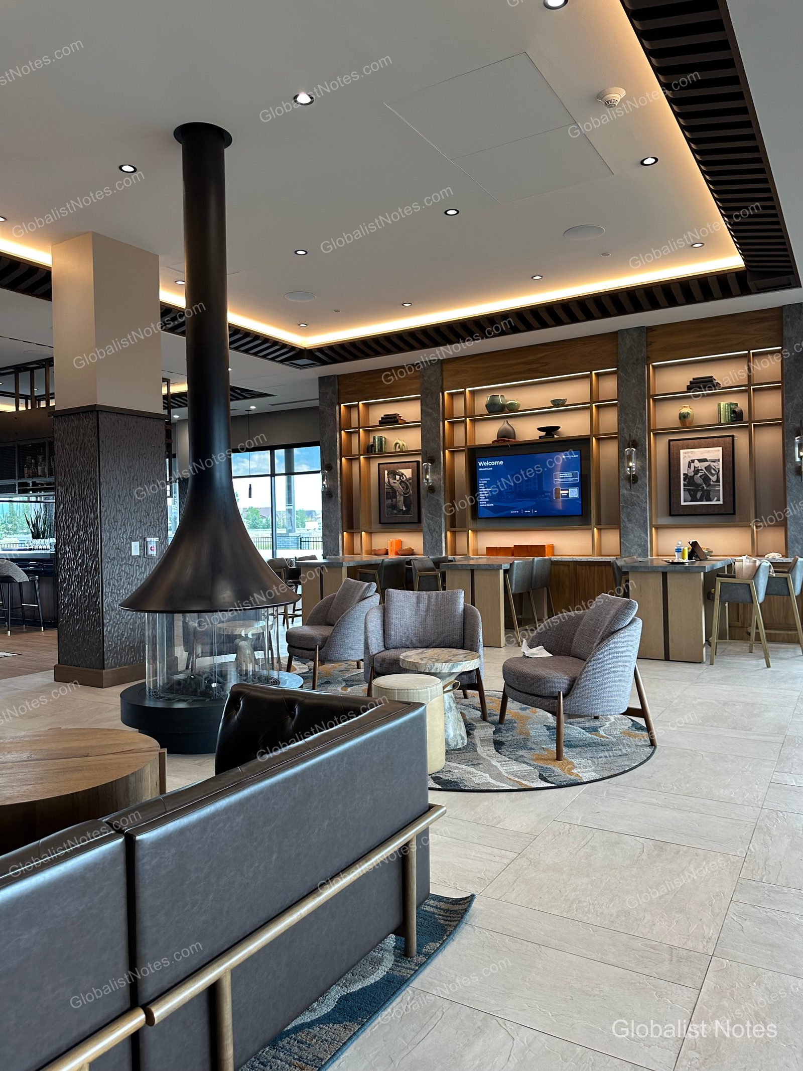 Hyatt Place Bozeman Yellowstone Airportのロビーラウンジ。中央に黒い円筒形の暖炉が配され、周囲にグレーのアームチェアと円形ラグが配置されています。壁面には照明付きの棚とテレビがあり、奥にはバーカウンターが見えます。