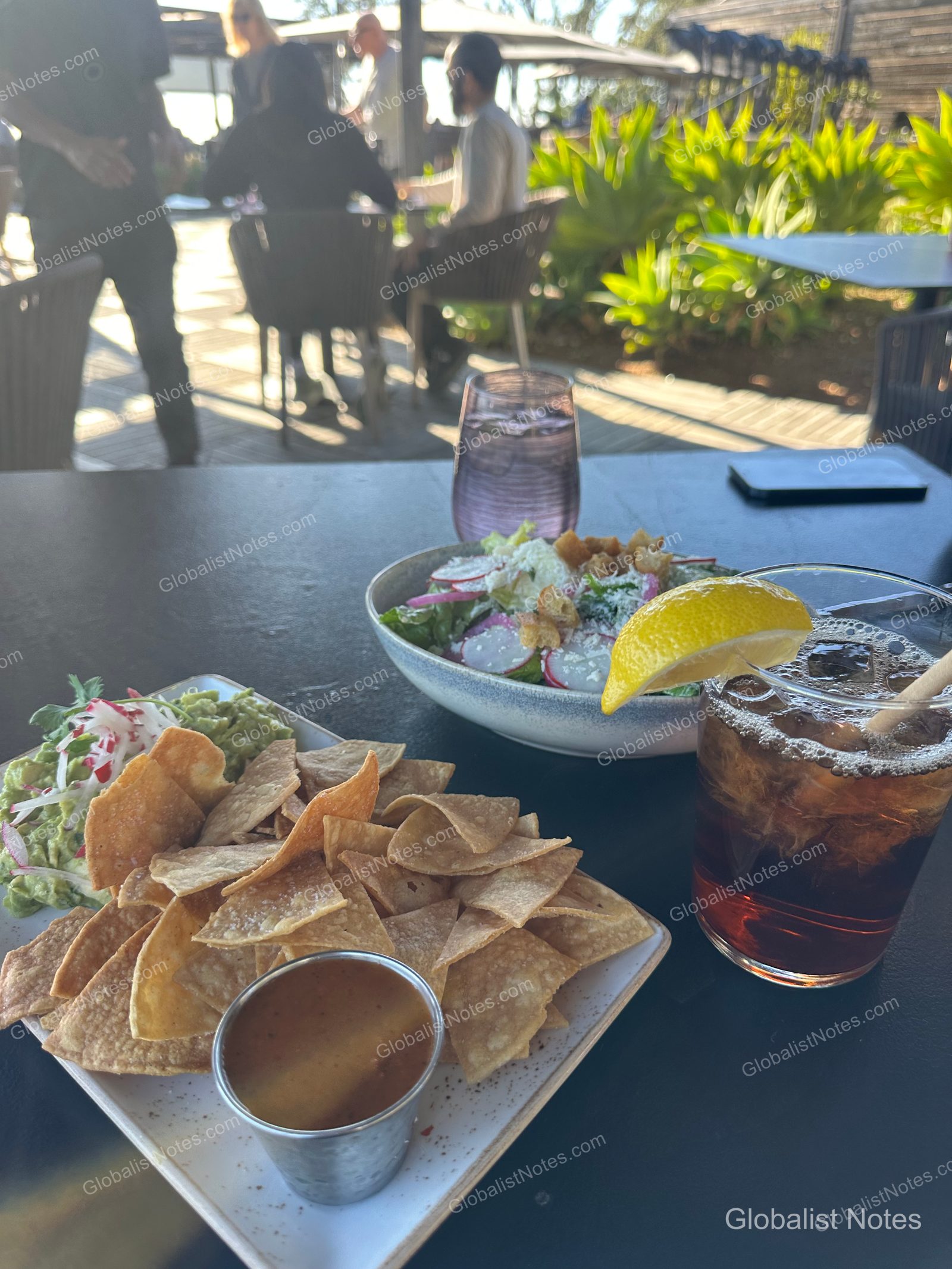Guacamole, nacho chips, a salad, and an iced drink with a lemon slice on an outdoor terrace table at Alila Ventana Big Sur, with other guests dining in the blurred background.