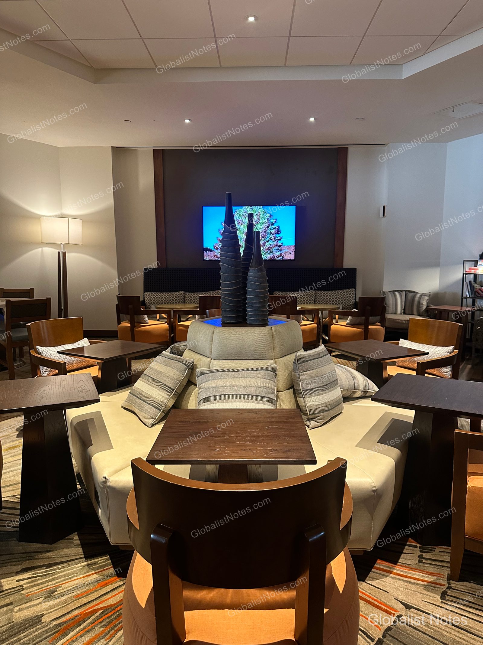Indoor lounge area at Hyatt Regency Maui Resort and Spa. The space features a central circular sofa with three tall decorative vases, surrounded by wooden tables and chairs. A large TV is mounted on the wall, creating a calm and orderly atmosphere.