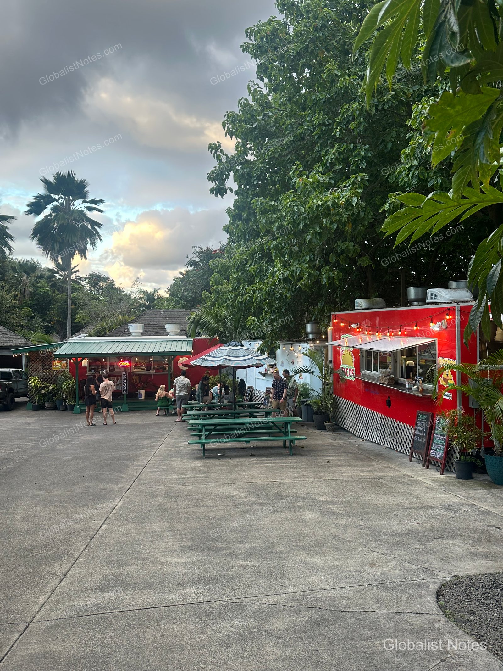 Outdoor area near Hana-Maui Resort featuring a row of red food trucks with wooden benches and umbrellas set up in front. Several people are ordering food or waiting nearby.