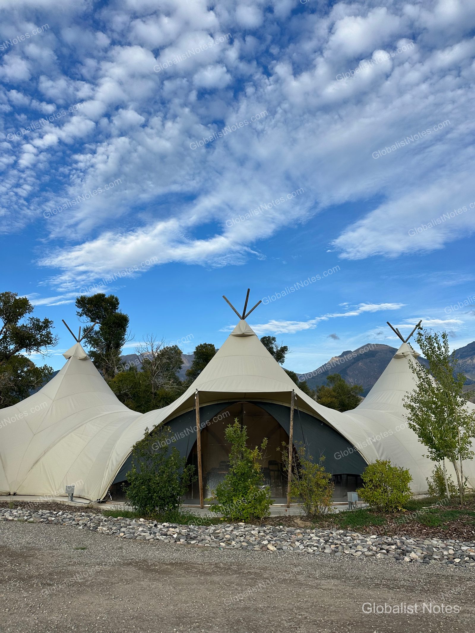 Under Canvas North Yellowstone - Paradise Valley（米国パインクリーク）の大型キャンバステント。3つの尖った屋根が連なる独特な構造で、手前には砂利道と木々、背景には山脈が広がっています。