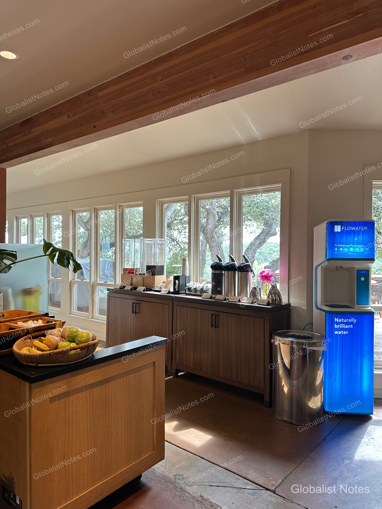 미라발 오스틴 리조트 앤 스파의 다이닝 공간에 마련된 셀프 서비스 스테이션입니다. 나무 수납장 위로 커피 서버와 음료 디스펜서가 놓여 있고, 옆에는 파란색 조명이 켜진 Flowater 정수기와 신선한 과일 바구니가 창가 쪽에 배치되어 있습니다.