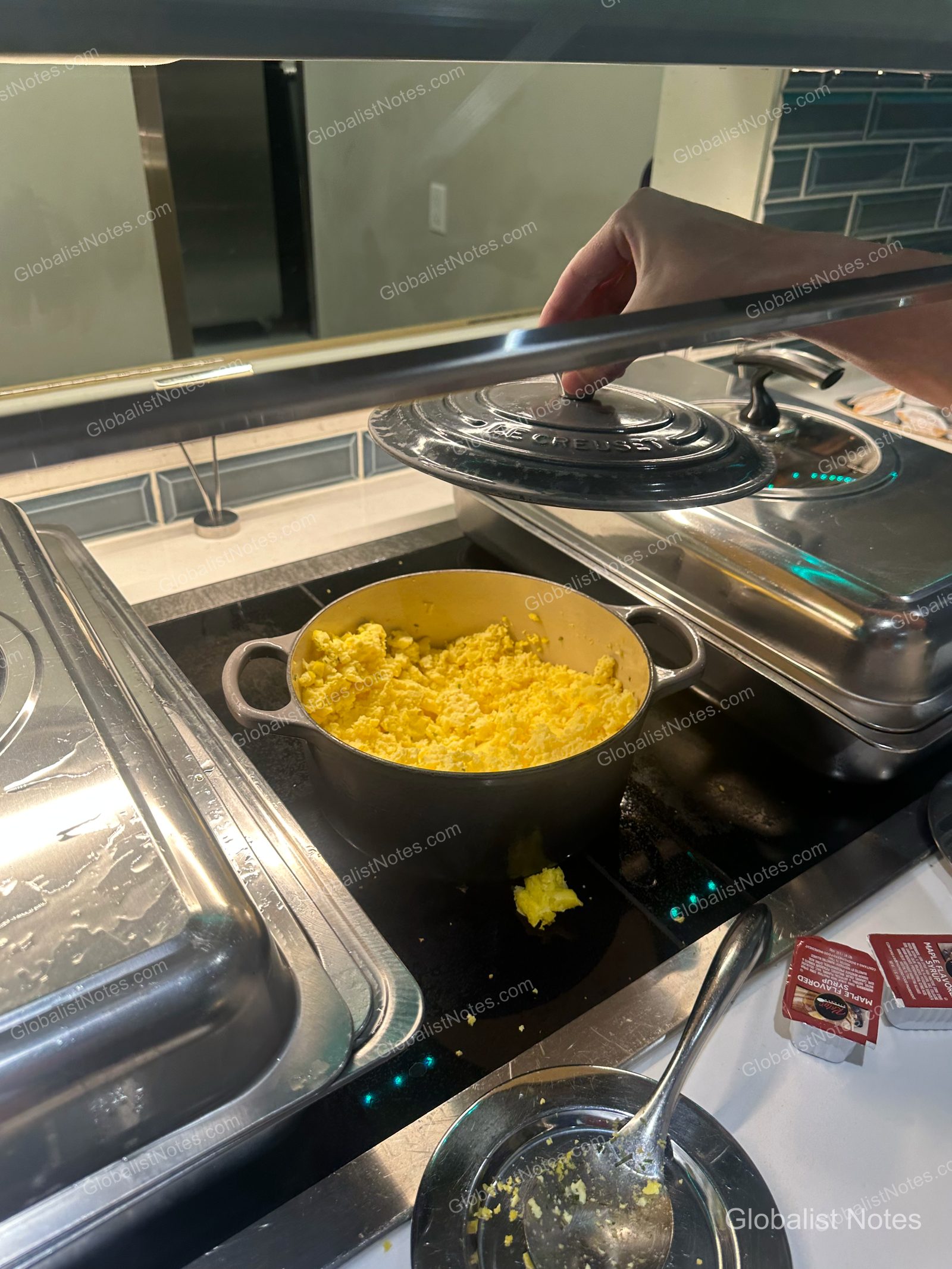 Hyatt Place Flagstaffの朝食ビュッフェ。ル・クルーゼの鍋の蓋を開け、スクランブルエッグを確認する様子。周囲にはビュッフェ用の容器やメープルシロップが並ぶ。