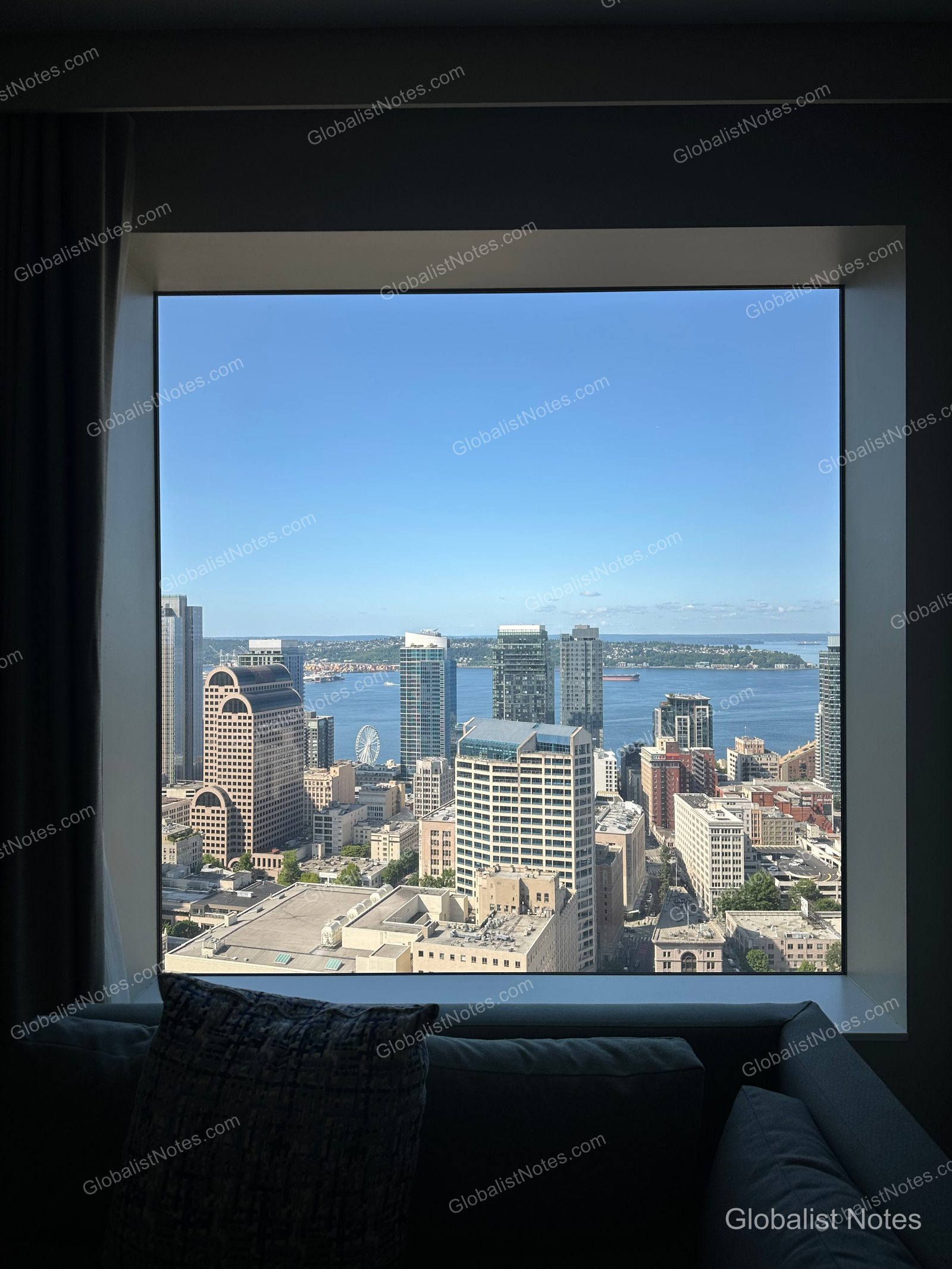 Daytime view from the floor-to-ceiling windows of a Hyatt Regency Seattle guestroom, overlooking the downtown skyline, the blue water in the distance, and the waterfront Ferris wheel under a clear sky.