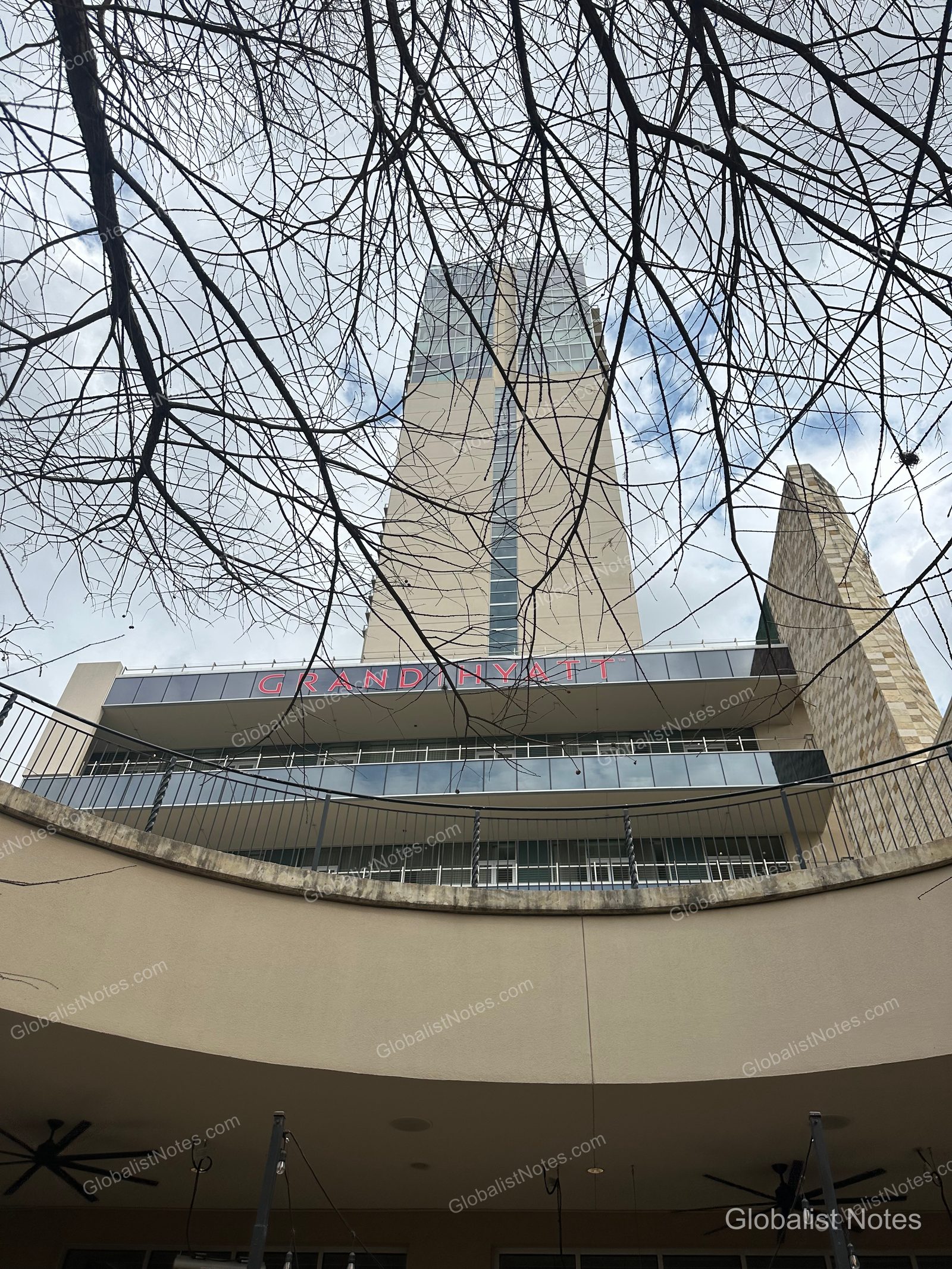 Exterior view of the Grand Hyatt San Antonio River Walk. The red Grand Hyatt logo is visible at the top of the modern high-rise building against a backdrop of bare tree branches.