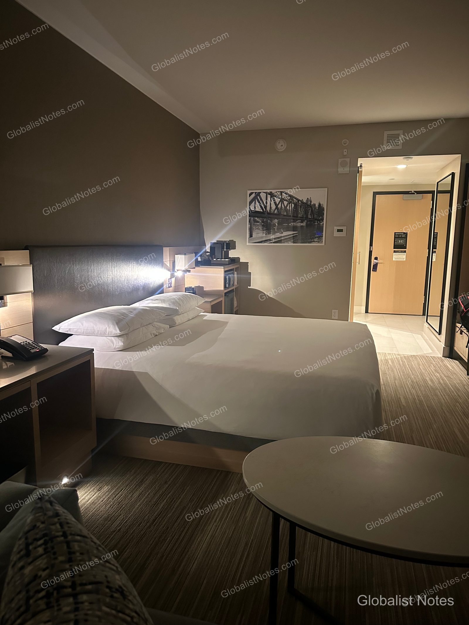 Interior of a Standard King room at Hyatt Regency Seattle. A king bed with white linens is centered under a framed photo of a Seattle steel bridge, with a coffee machine on the nightstand. To the right, the room entrance, hallway, and a full-length mirror are visible.