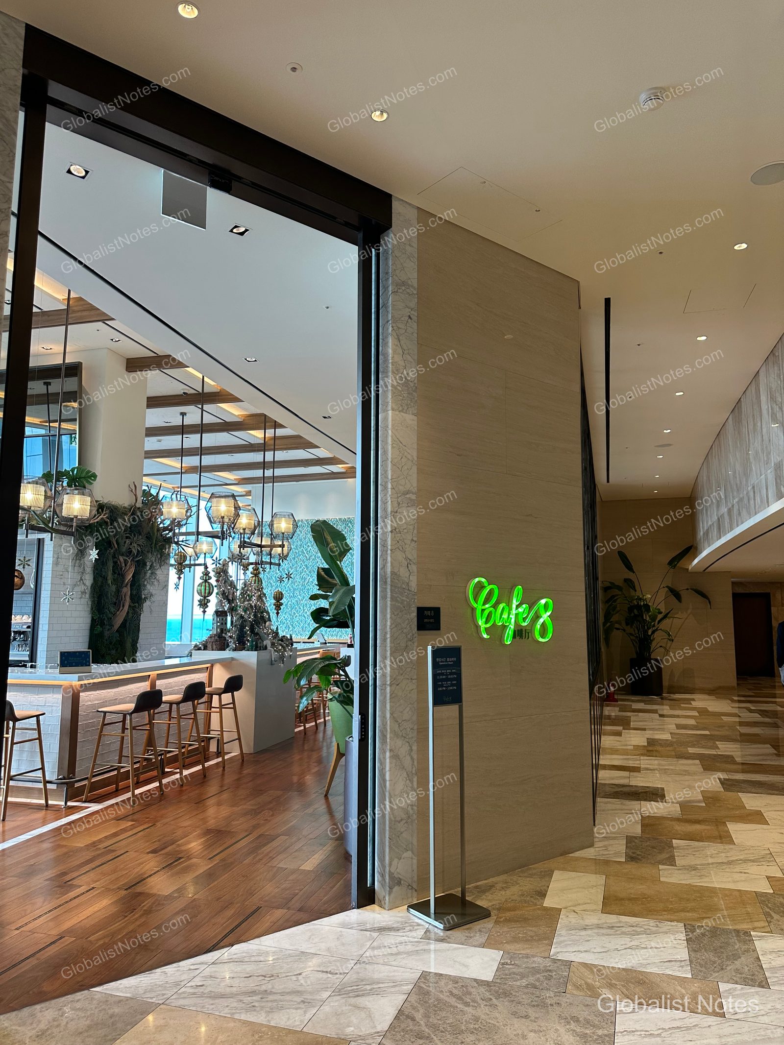 Entrance to Cafe 8 at Grand Hyatt Jeju. A bright lime green neon sign is mounted on the wall, with a view into the interior featuring a bar counter, pendant lighting, wooden flooring, and light marble-patterned floors.