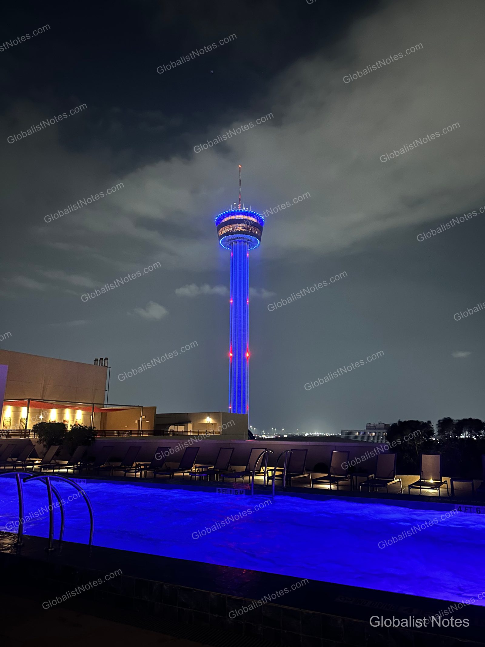 The Tower of the Americas illuminated against the night sky, viewed from the outdoor pool area of Grand Hyatt San Antonio River Walk, which features blue accent lighting and lounge chairs.