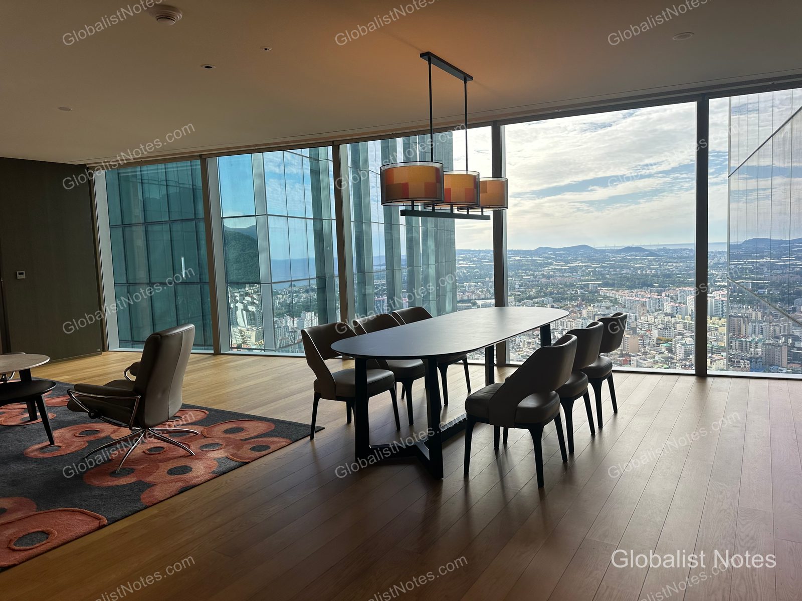 Living area of a One Bedroom Suite at Grand Hyatt Jeju. Floor-to-ceiling windows offer a view of Jeju City, with a six-person dining table and chairs in the center, and a comfortable sofa with a patterned rug on the left.