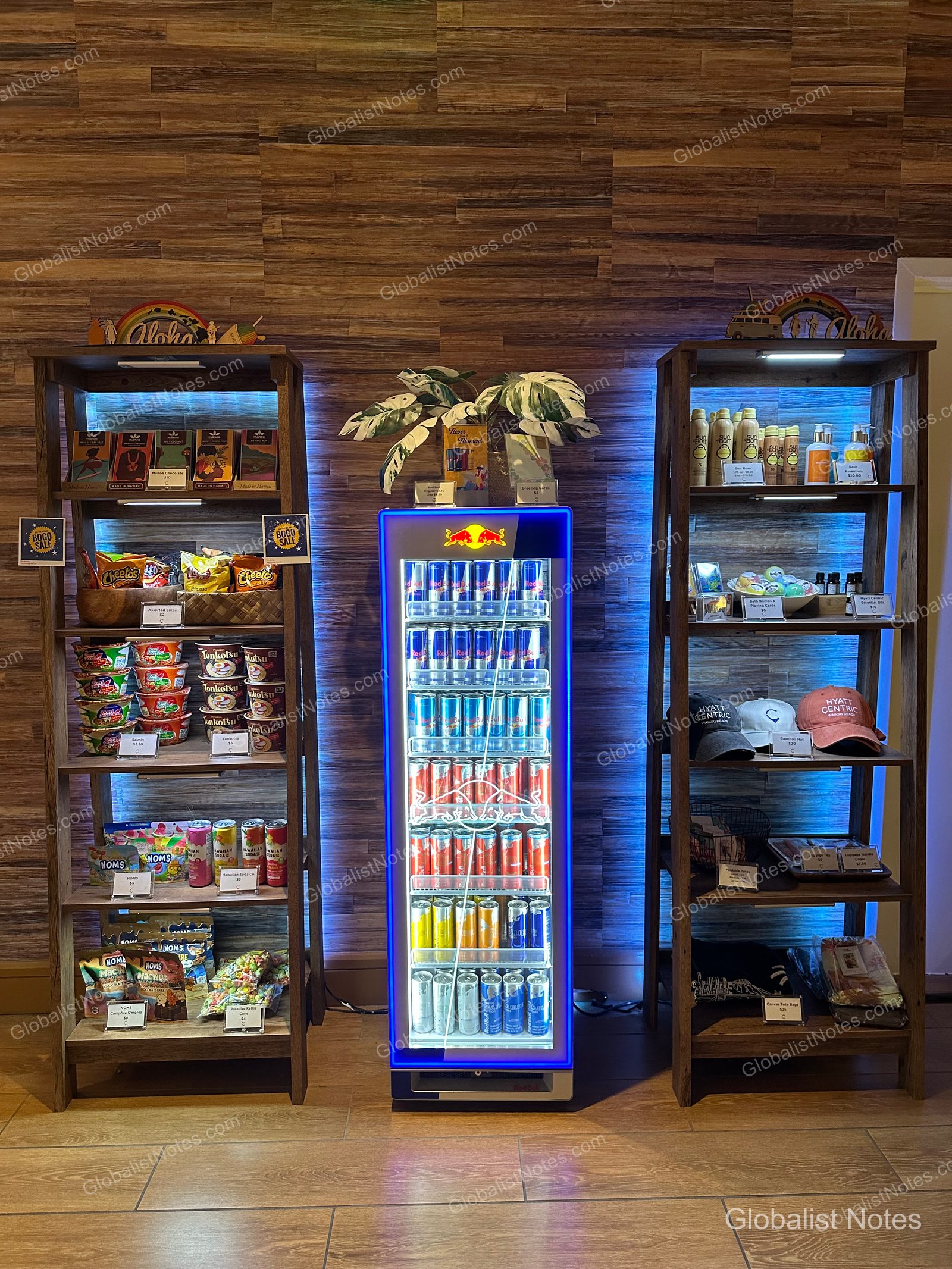 The lobby sundry shop at Hyatt Centric Waikiki Beach. Wooden shelves on either side display snacks, cup noodles, hats, and sun care products, while a blue-lit Red Bull refrigerator is positioned in the center.