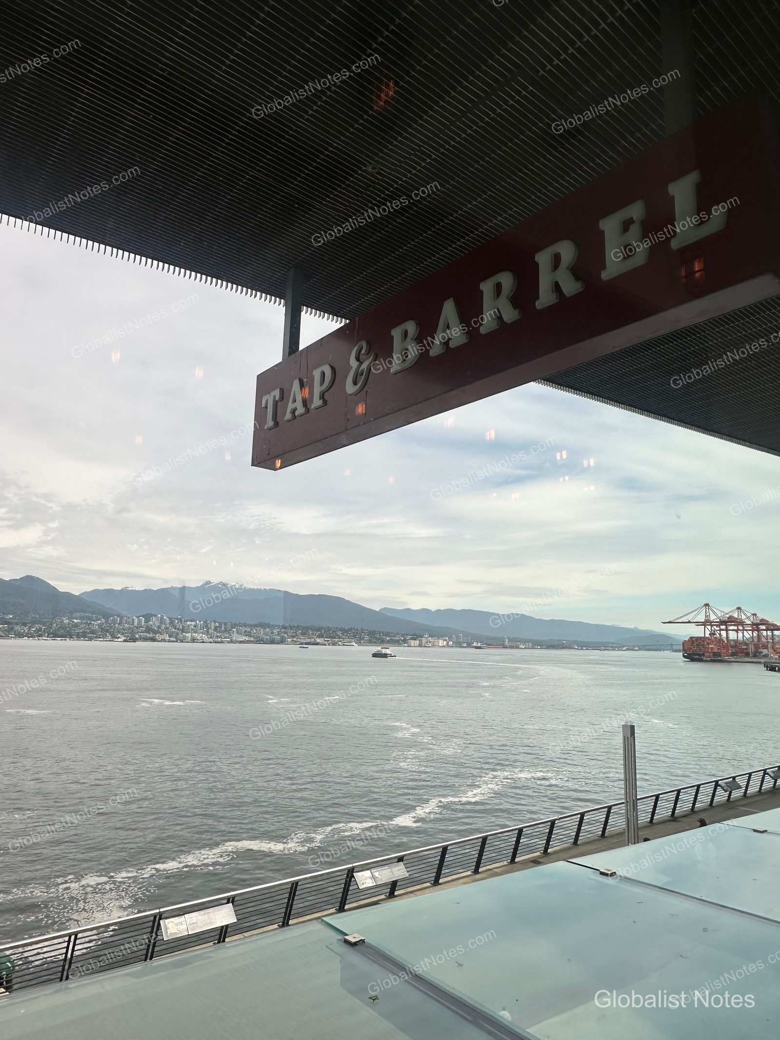 View of the harbor, distant mountains, and port cranes from beneath the Tap & Barrel restaurant sign near Canada Place in Vancouver. The scene looks out over a glass railing toward the calm water under a cloudy sky.
