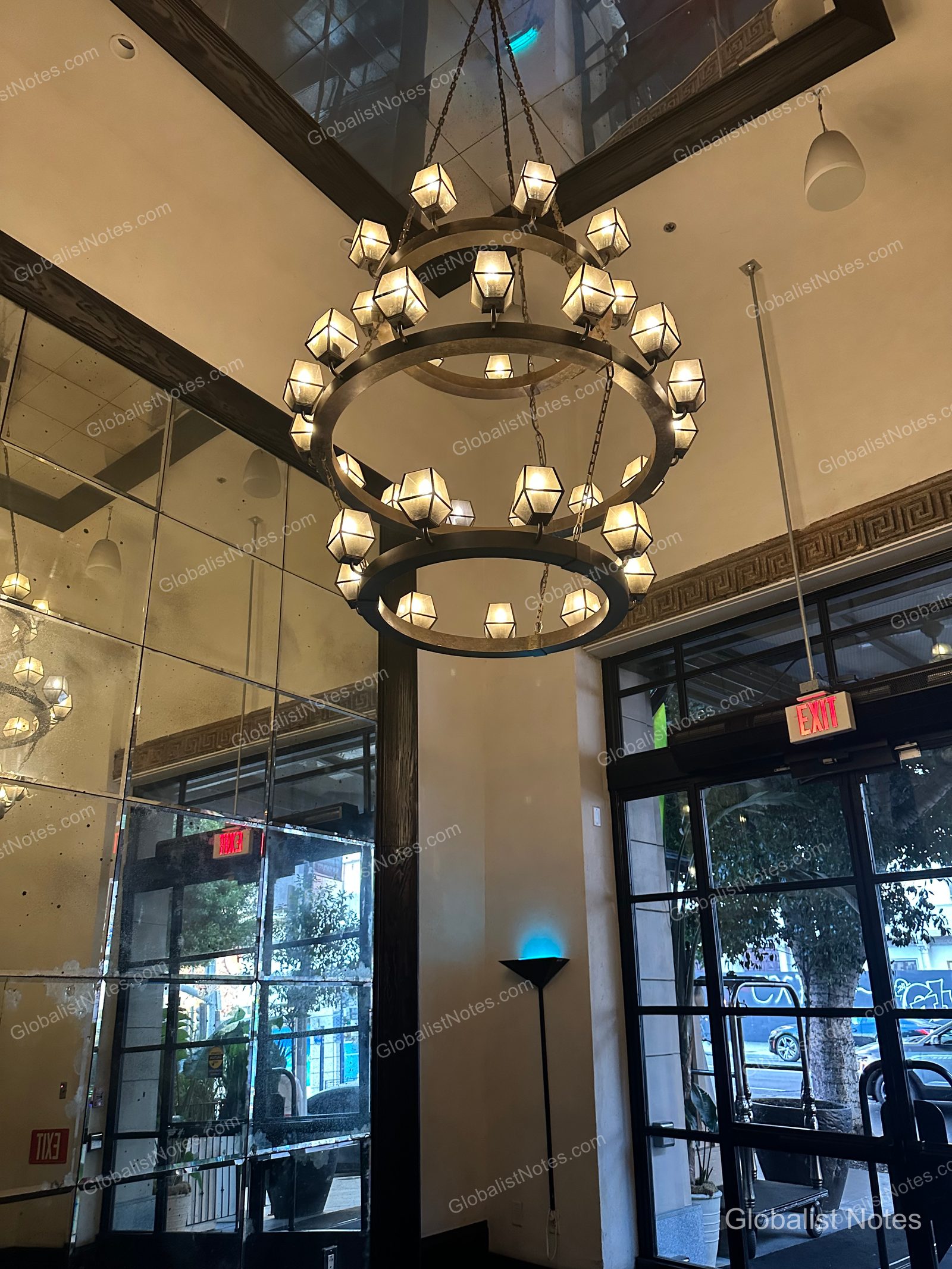 Interior view of the Hotel Figueroa Los Angeles lobby, featuring a two-tier circular metal chandelier hanging from the ceiling, a long vintage mirrored wall, and a view of the street through the windows.
