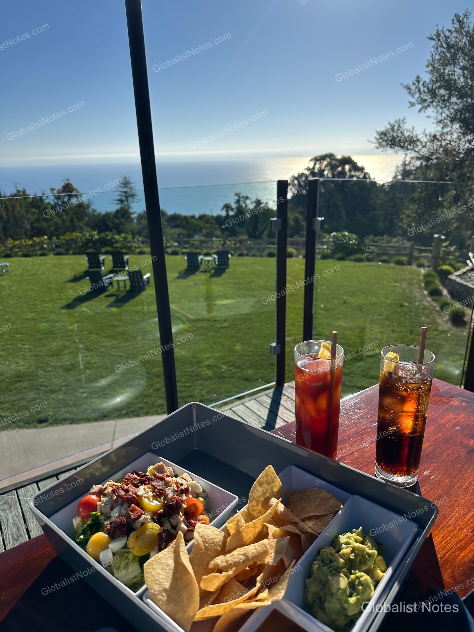 Salad, nachos with guacamole, and two drinks on an outdoor terrace table at Alila Ventana Big Sur. In the background, the sunlit lawn and the Pacific Ocean are visible beyond the glass railing.