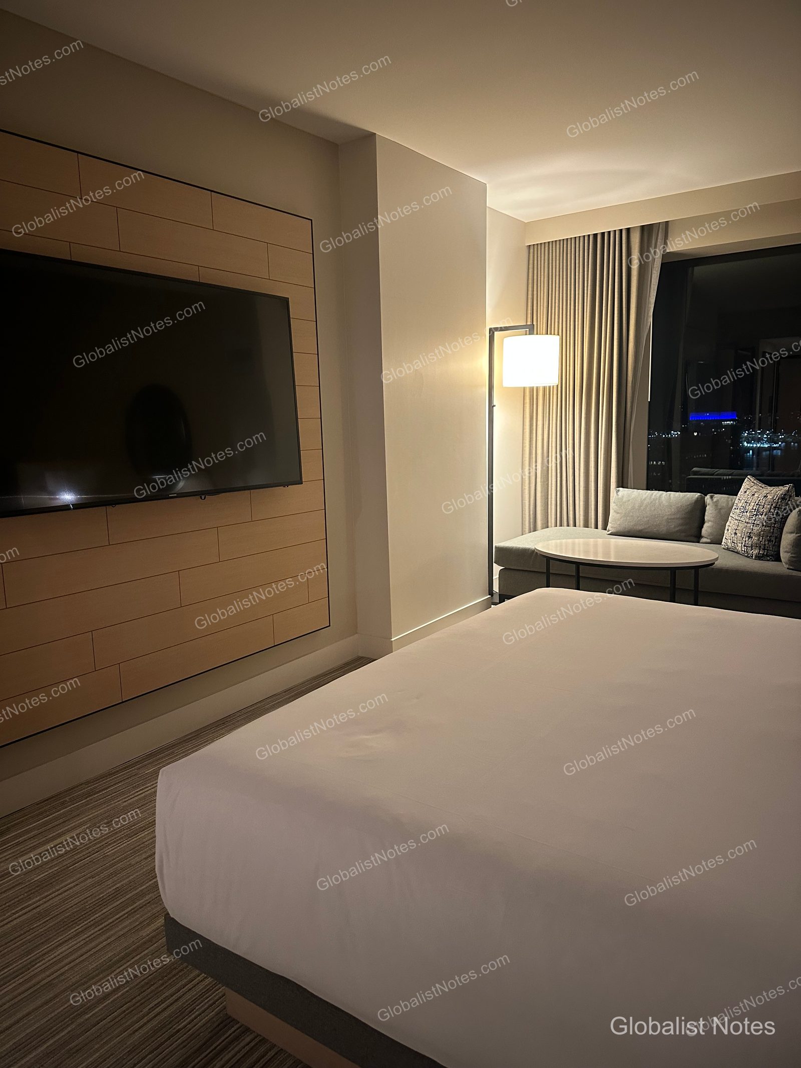 Interior of a Standard King room at Hyatt Regency Seattle. A king bed and wall-mounted TV are positioned in the room, with a sofa, round table, and floor lamp by the window. The window overlooks the Seattle city skyline at night.