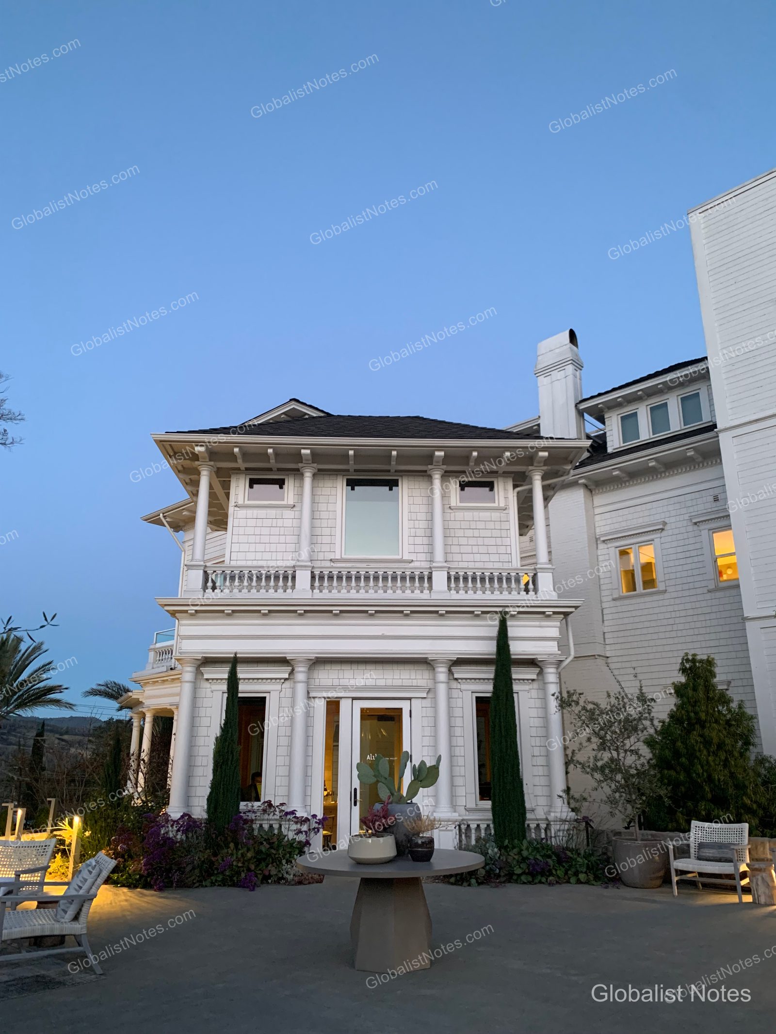 Exterior view of Alila Napa Valley in St. Helena, USA, featuring white wooden buildings with round tables and chairs set in the garden. Tall trees flank the entrance against a twilight blue sky, with outdoor furniture on the terrace.