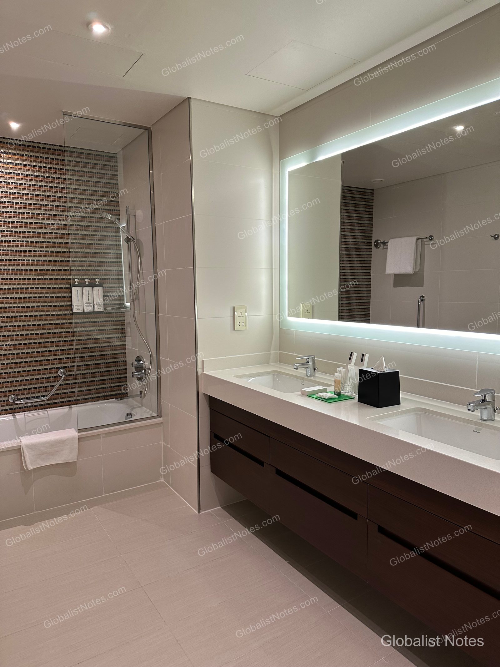 Hyatt Place Dubai Wasl District 一房套房浴室。左側為棕色馬賽克磁磚牆面的浴缸與淋浴設備，右側為配有大型背光鏡面與雙洗手台的長型檯面。