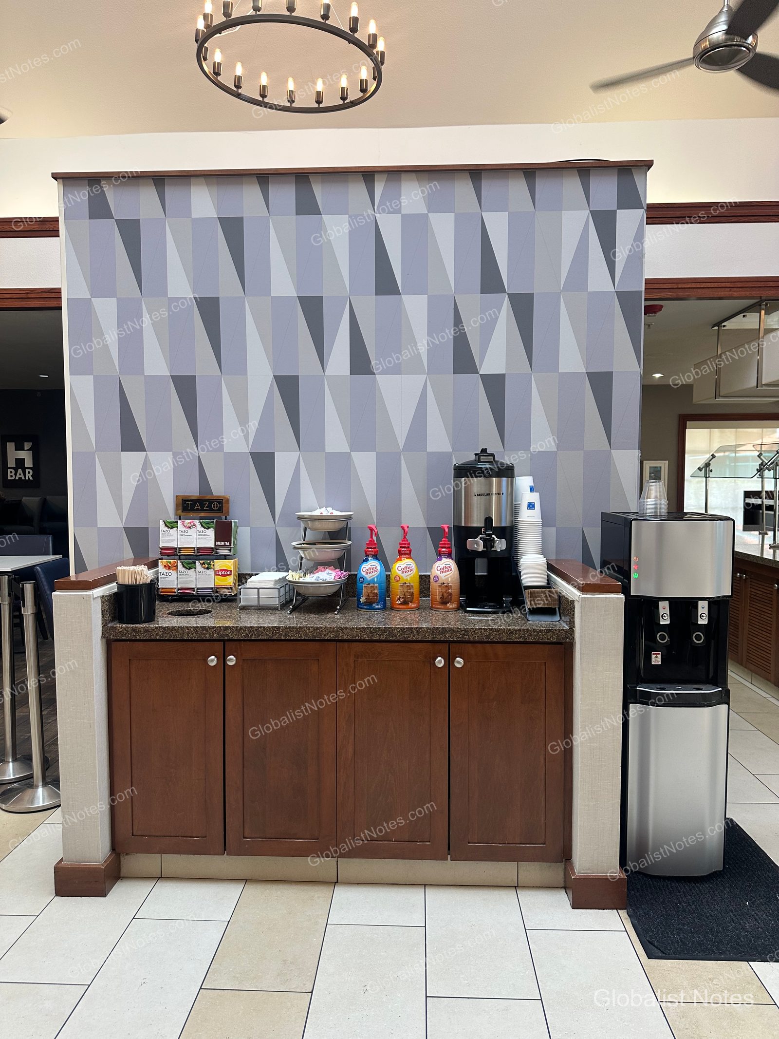 Hyatt House Scottsdale/Old Townの朝食エリアのドリンクステーション。ブラウンの棚にTazoのティーバッグ、コーヒーマシン、コーヒーメイト、浄水器が並び、背面の壁はグレーの幾何学模様で装飾されています。