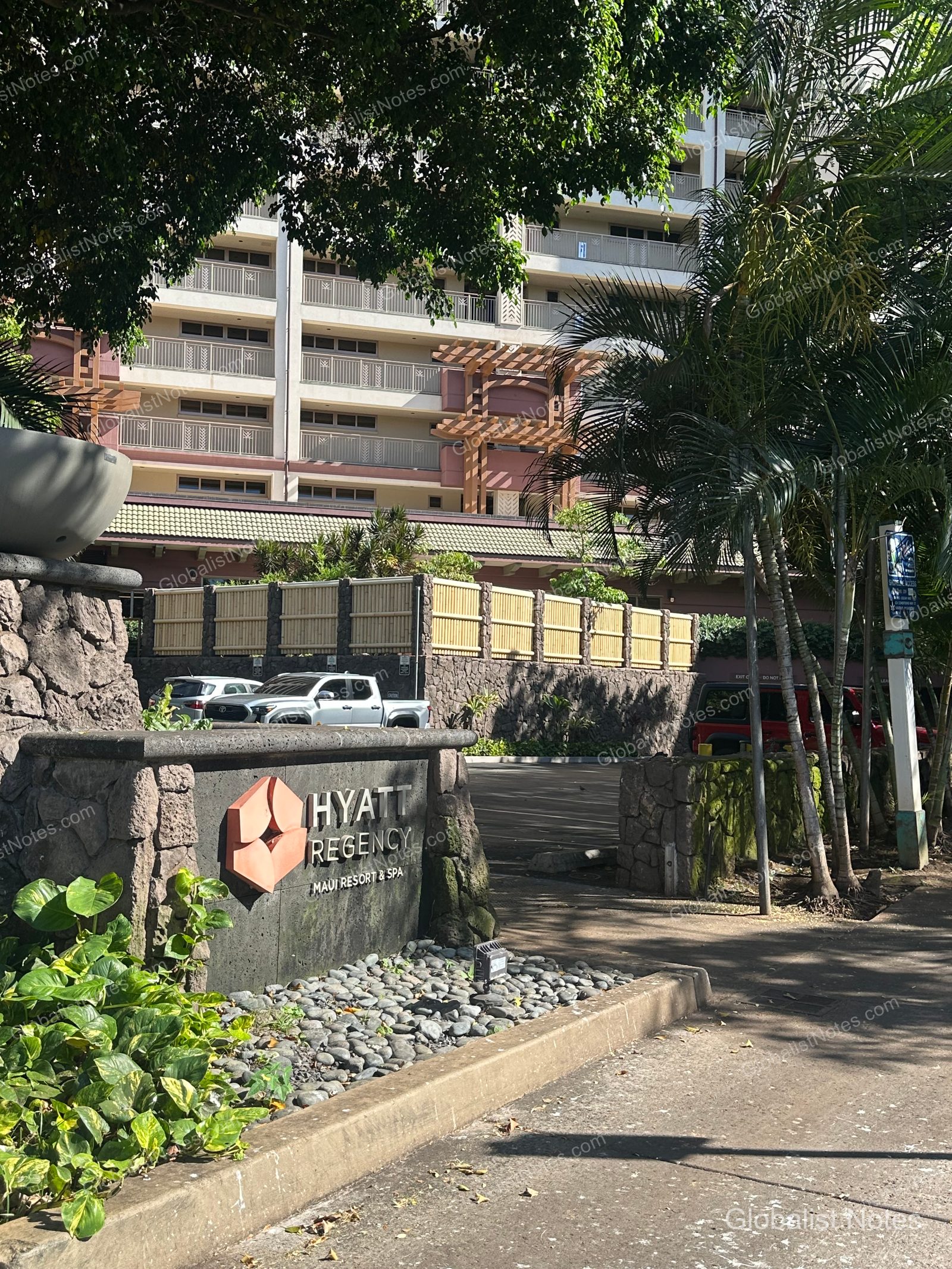 Entrance sign for Hyatt Regency Maui Resort and Spa in Lahaina, Hawaii, with the resort building in the background. The stone-finished sign is surrounded by palm trees and greenery, with the road leading to the parking area and a portion of the multi-story resort building visible in the sunlight.